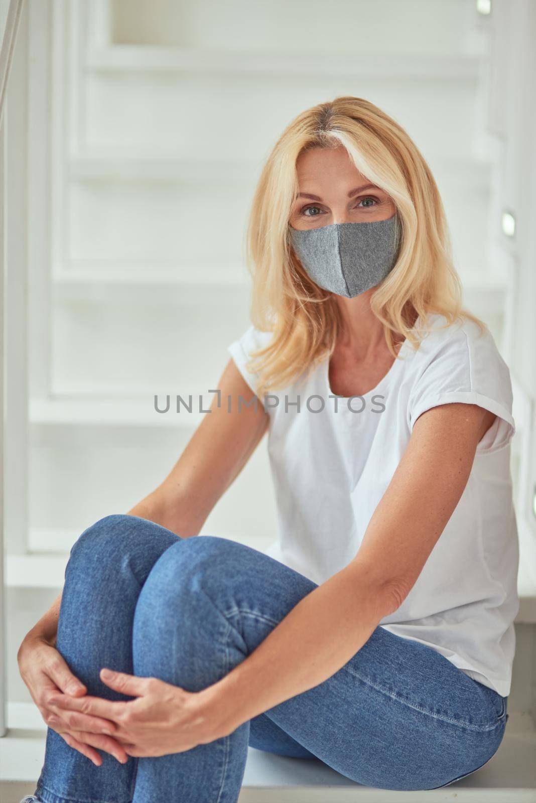 Middle aged caucasian woman wearing protective face mask during coronavirus pandemic sitting on white stairs at home. Isolation at home concept
