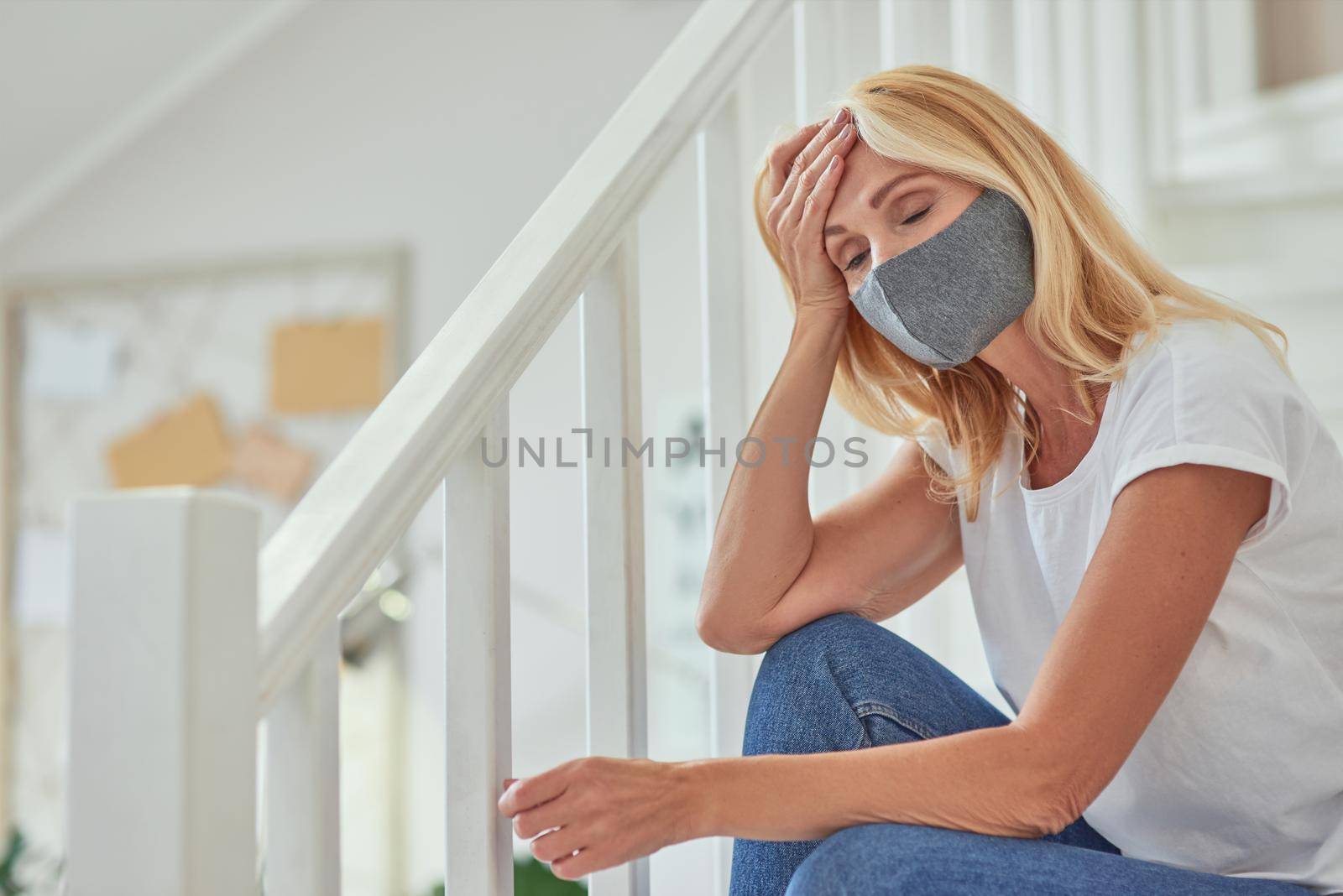 Sick middle aged caucasian woman wearing protective face mask sitting on stairs in light modern flat. Isolation at home concept