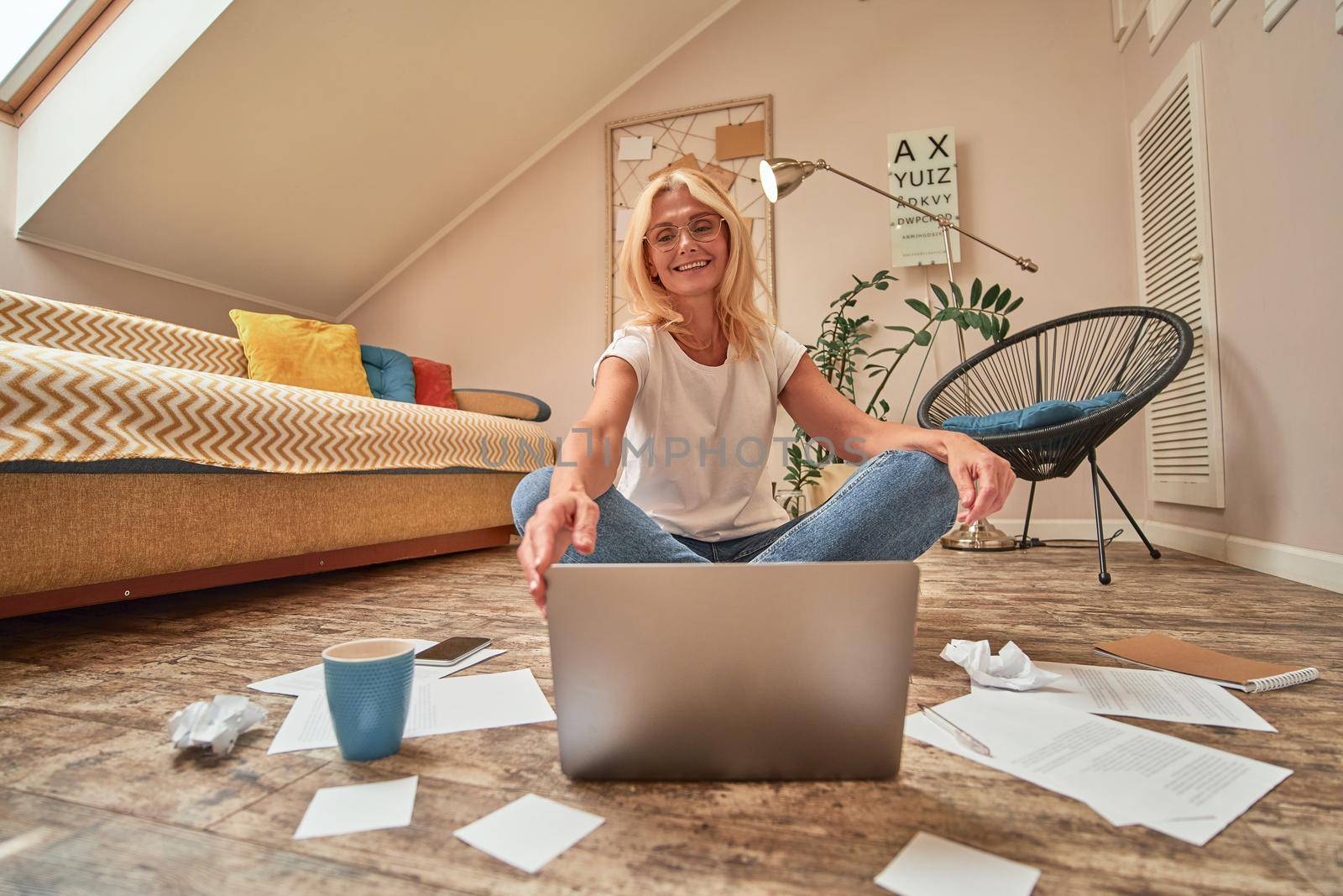 Smiling middle aged caucasian woman working on floor with laptop and papers in modern apartment. Working from home concept