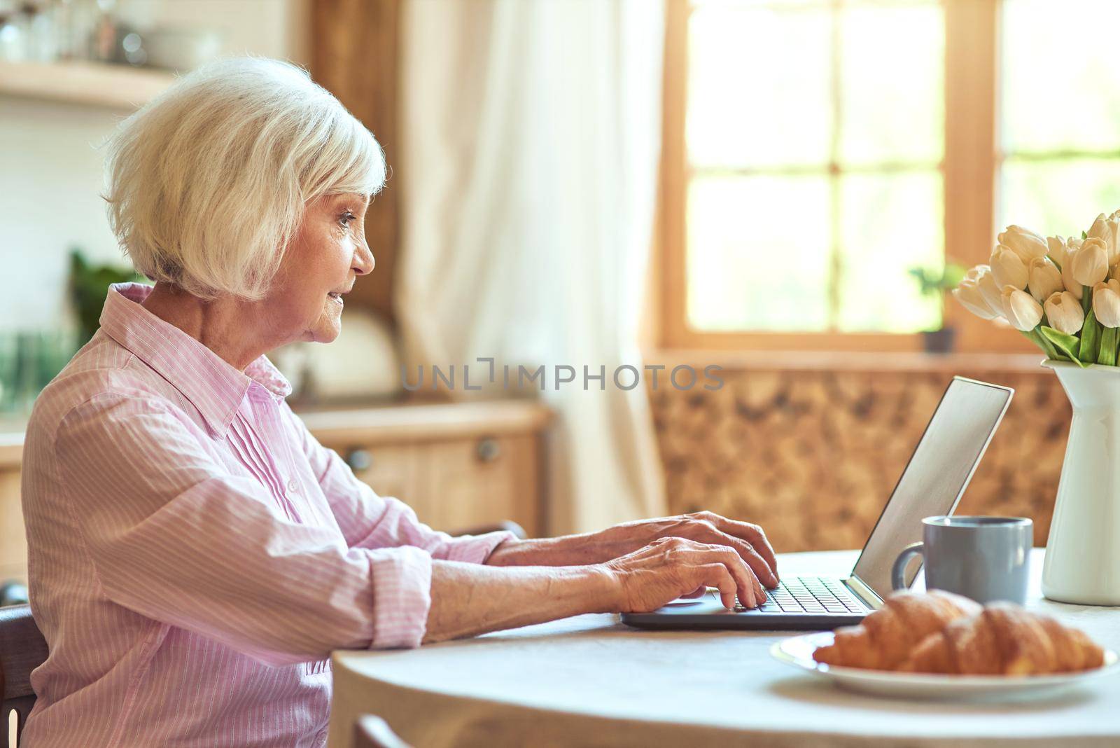 Side view of aged woman working at laptop while drinking tea during morning at home. Domestic lifestyle concept. Copy space