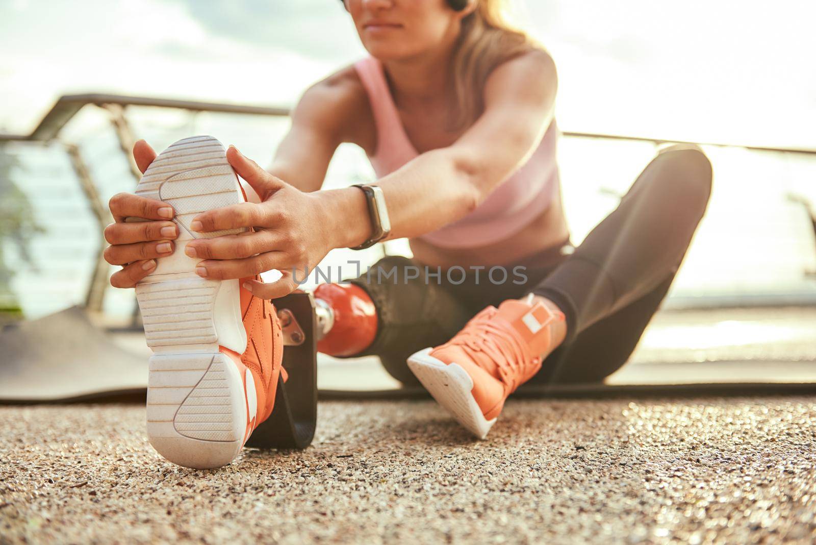 Stretching exercises. Cropped photo of young beautiful woman in headphones with leg prosthesis listening music and stretching her leg while sitting on the bridge by friendsstock