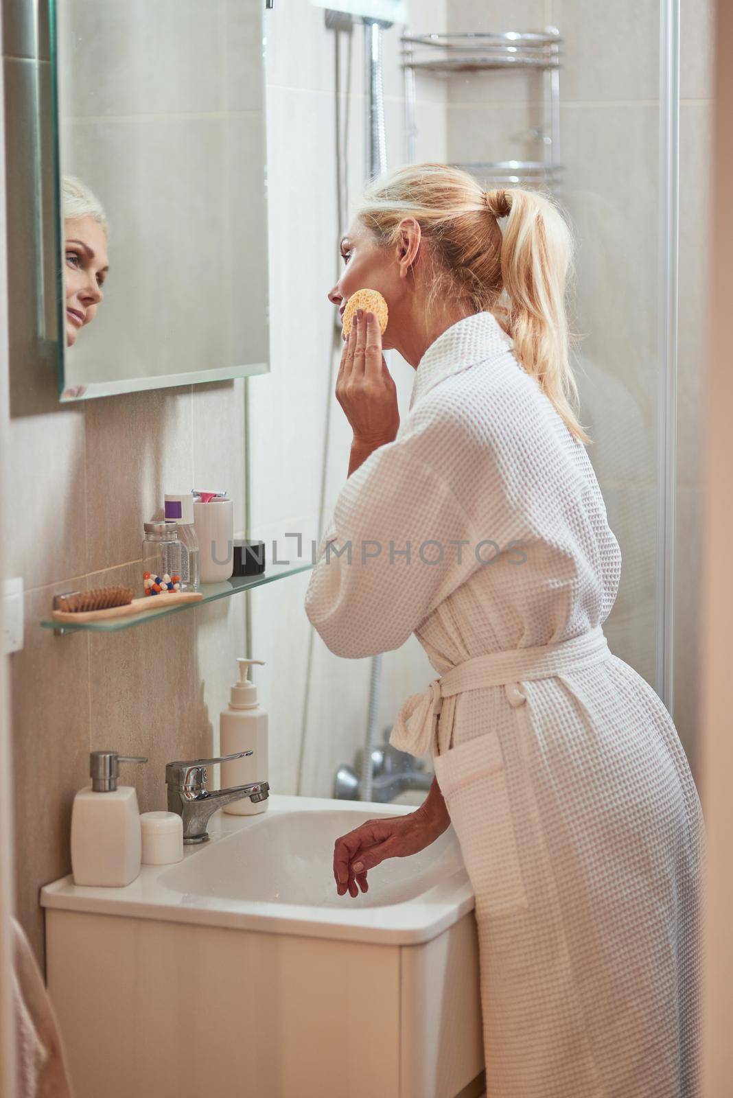 Smiling middle aged caucasian woman cleaning face with sponge standing in front of bathroom mirror. Beauty, skincare and cosmetology concept