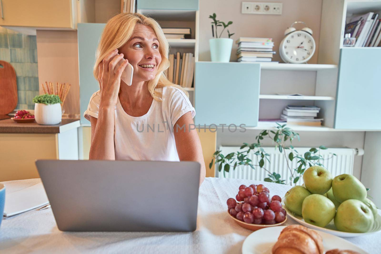 Happy middle aged caucasian woman talking on phone while sitting on home kitchen near laptop and looking sideways. Working from home concept