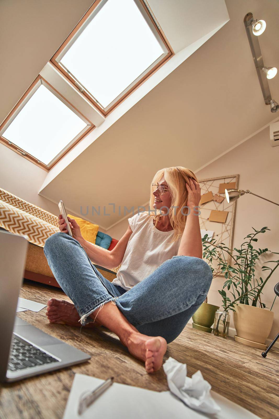 Middle aged caucasian woman sitting next to laptop on light flat floor with crossed legs and looking into smartphone. Working from home concept