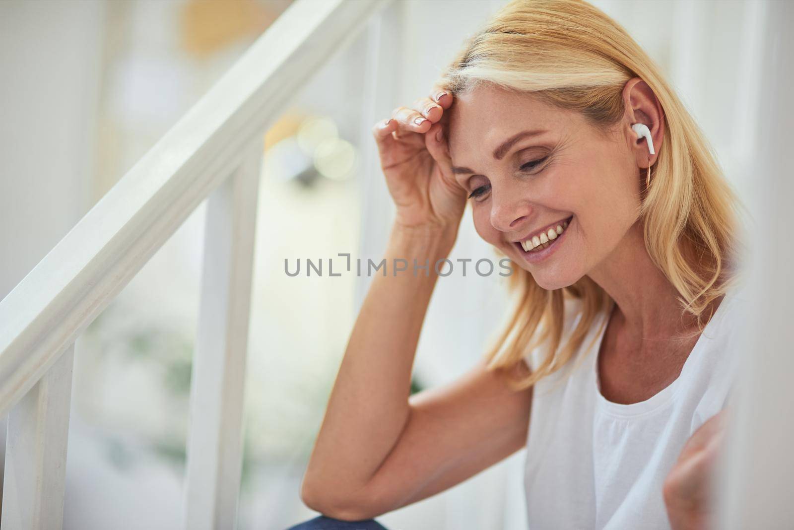 Pretty middle aged caucasian woman with wireless earphone looking downwards and smiling indoors. People spending time at home