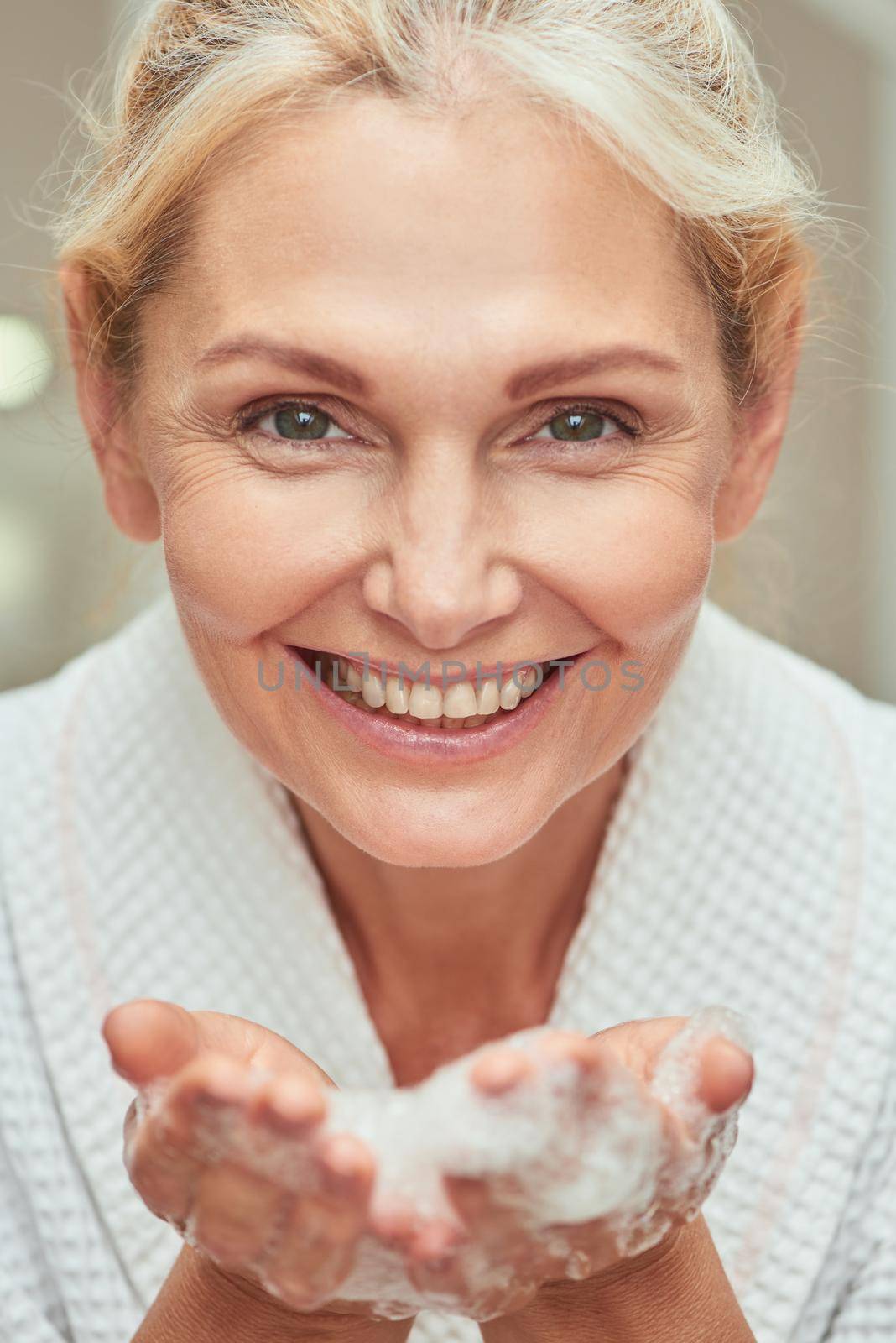 Close up portrait of happy middle aged caucasian woman with open palms full of foam looking at camera and smiling at spa. Beauty, skincare and cosmetology concept