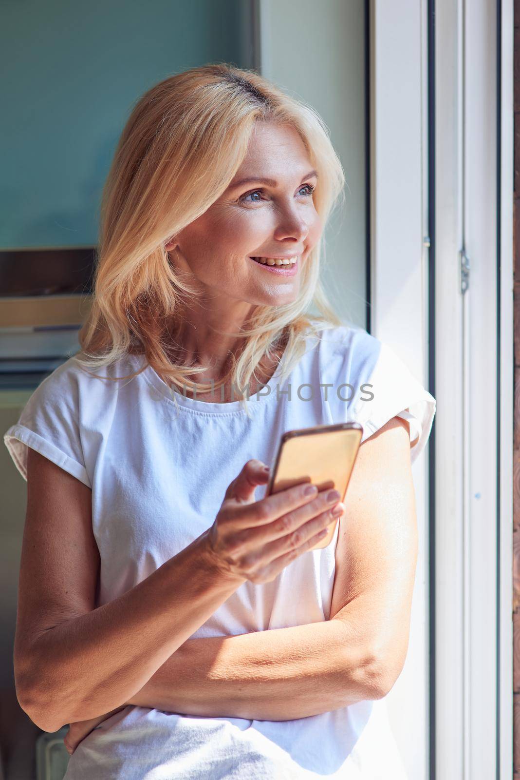 Middle aged caucasian woman holding phone while standing near sunny balcony and looking aside. People at home