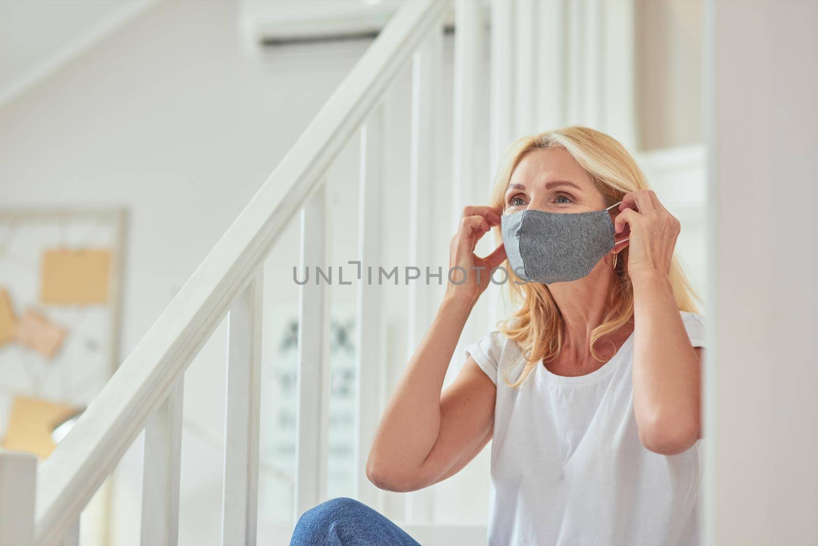 Middle aged caucasian woman putting protective mask on face sitting at modern apartment alone. Isolation at home concept