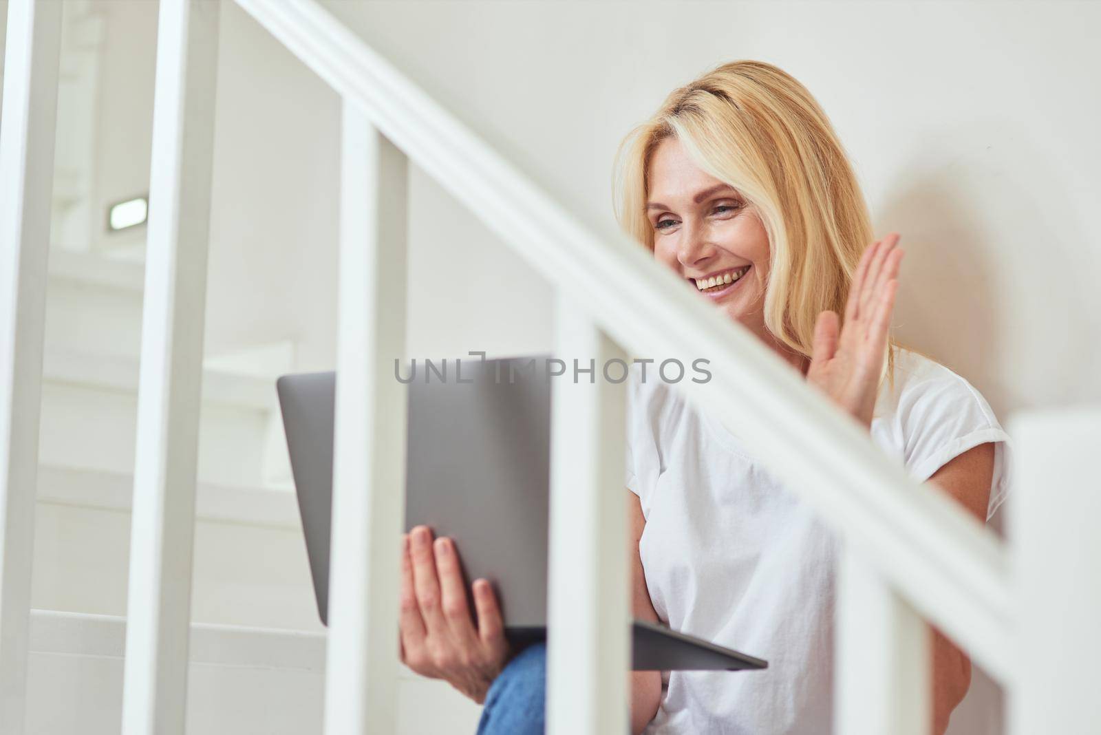 Happy middle aged caucasian woman talking via webcam on laptop and waving hand while sitting on stairs in modern flat. Freelance, working from home