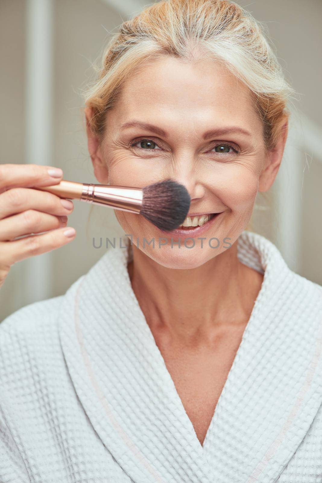 Beautiful middle aged caucasian woman looking at camera and smiling while applying makeup brush on face. Beauty, skincare and cosmetology concept