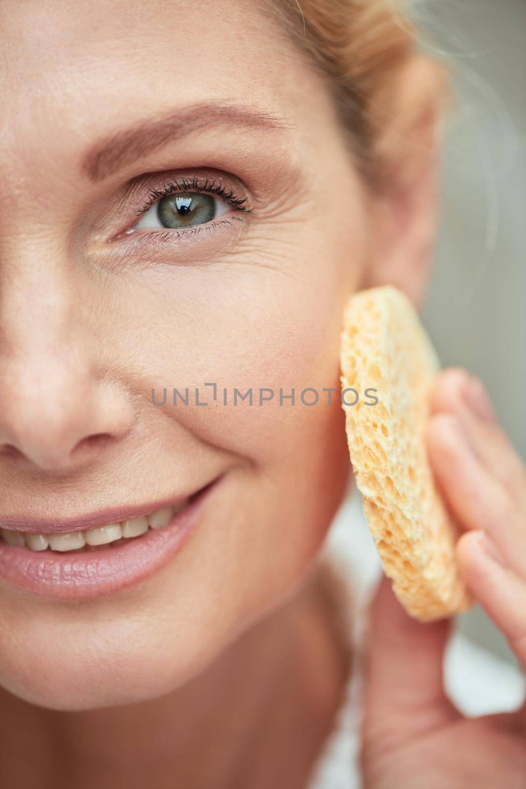 Beautiful middle aged caucasian woman with clean shiny skin holding facial sponge while looking at camera and smiling. Beauty, skincare and cosmetology concept