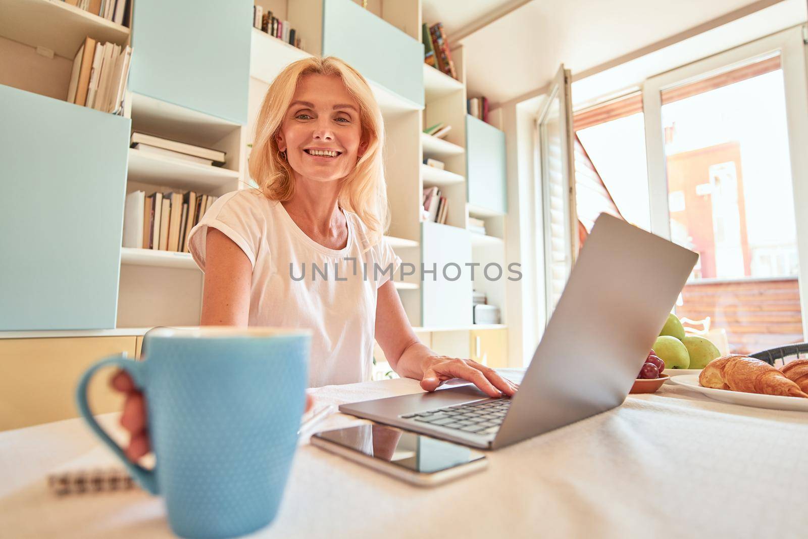 Middle aged caucasian business woman typing on laptop while taking cup of coffee from kitchen table in light apartment, selective focus. Working from home concept