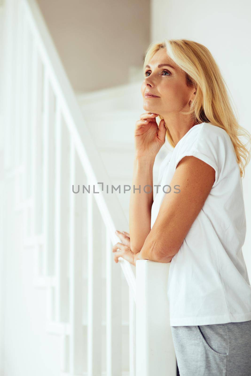 Portrait of pretty middle aged caucasian woman standing sideways looking up on stairs indoors. People spending time at home