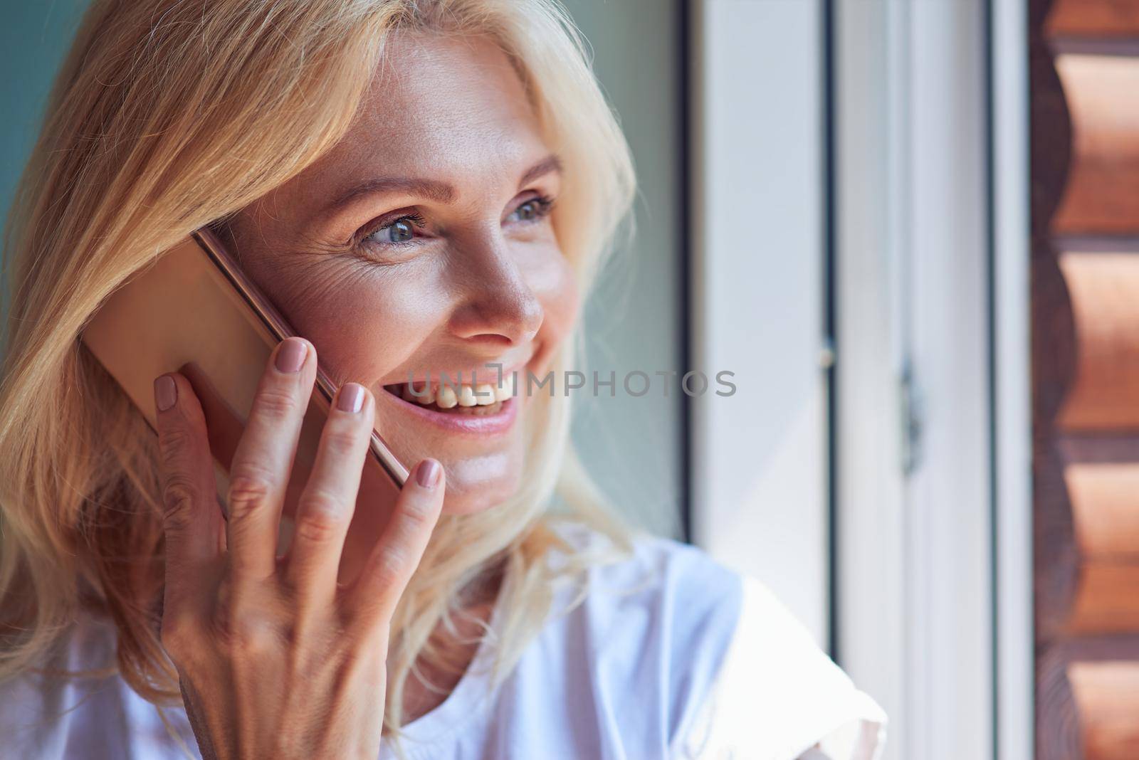 Portrait of middle aged caucasian woman looking outside to balcony standing indoors with phone near ear. People spending time at home