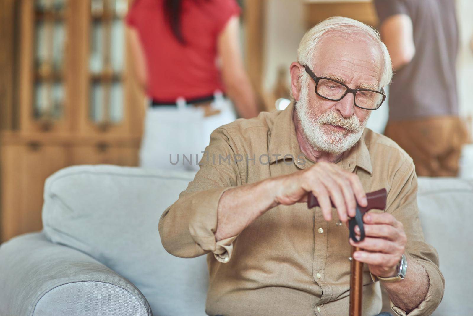 Waist up of senior man sitting on sofa and holding walking stick with people on the background. Care and health concept. Copy space
