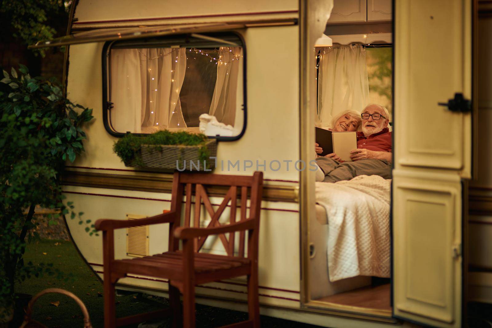 Happy senior married couple lying on the couch in their motorhome and reading a book in the evening. Travel concept