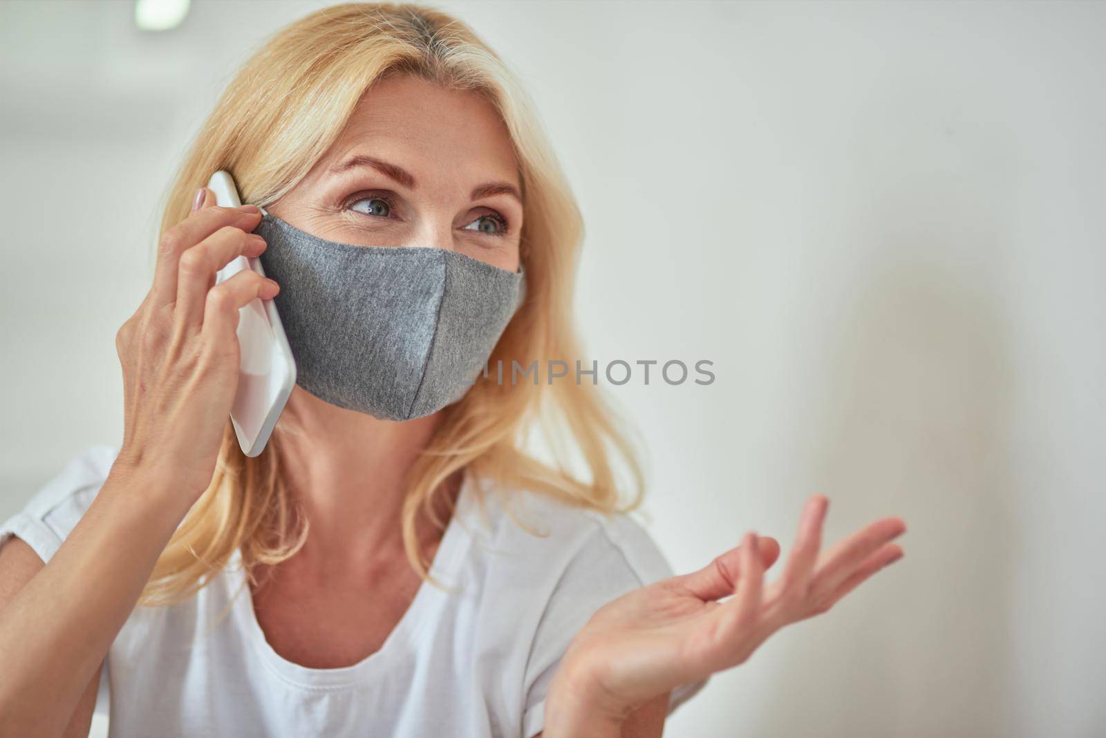 Middle aged caucasian woman with protective mask looking sideways and gesturing while talking on phone sitting at home. Isolation at home concept