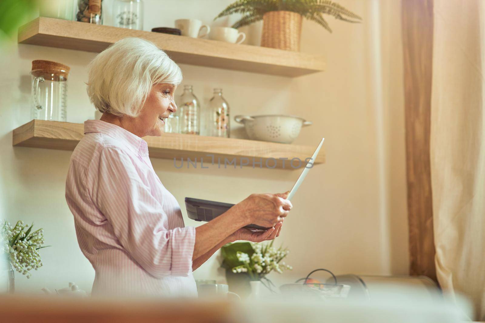 Side view of senior female standing and holding laptop while looking at screen at the kitchen. Domestic lifestyle concept