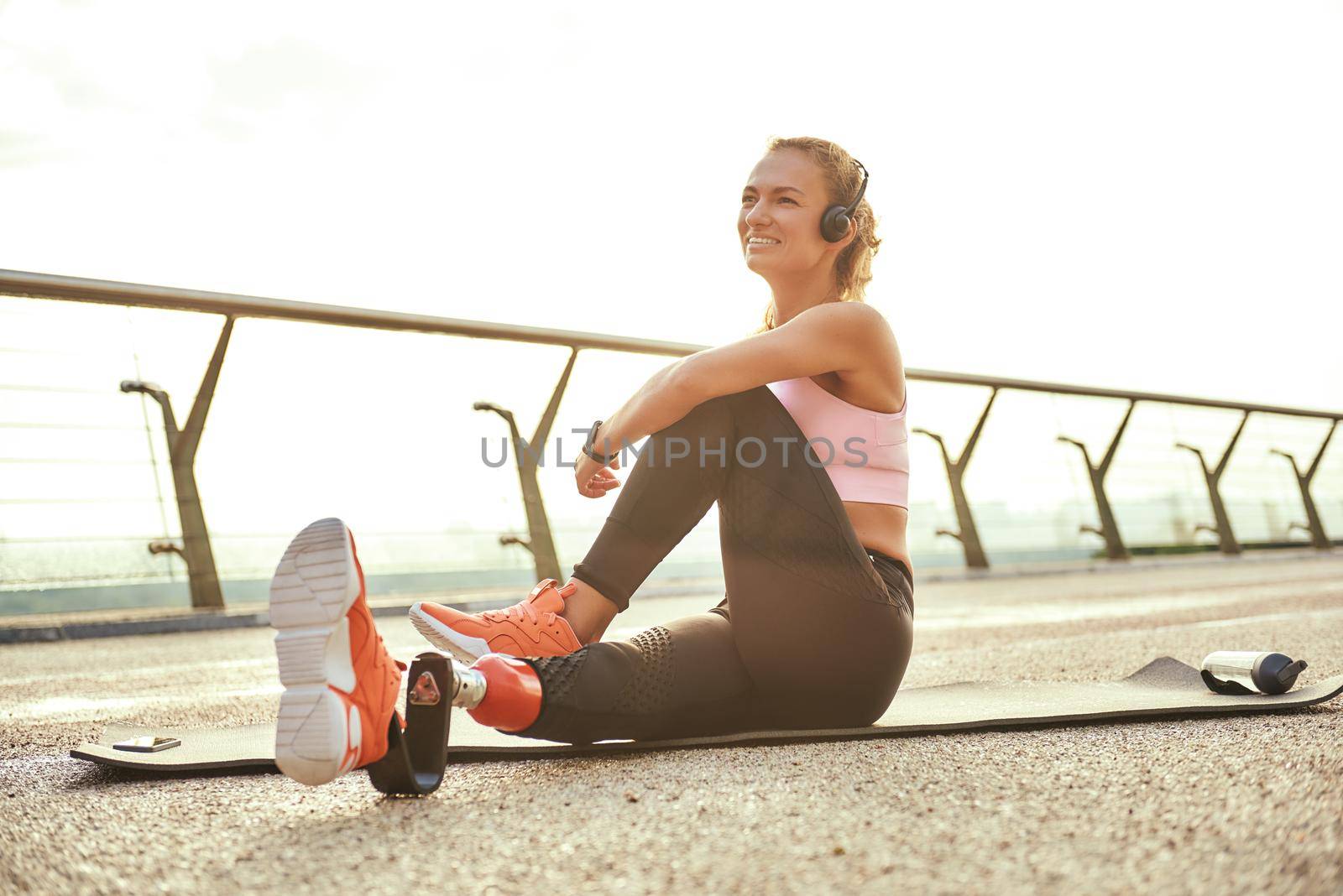 Happy morning. Positive disabled athlete woman with prosthetic leg in headphones doing yoga exercises and smiling while sitting on the bridge. Disabled sport concept. Motivation. Healthy lifestyle