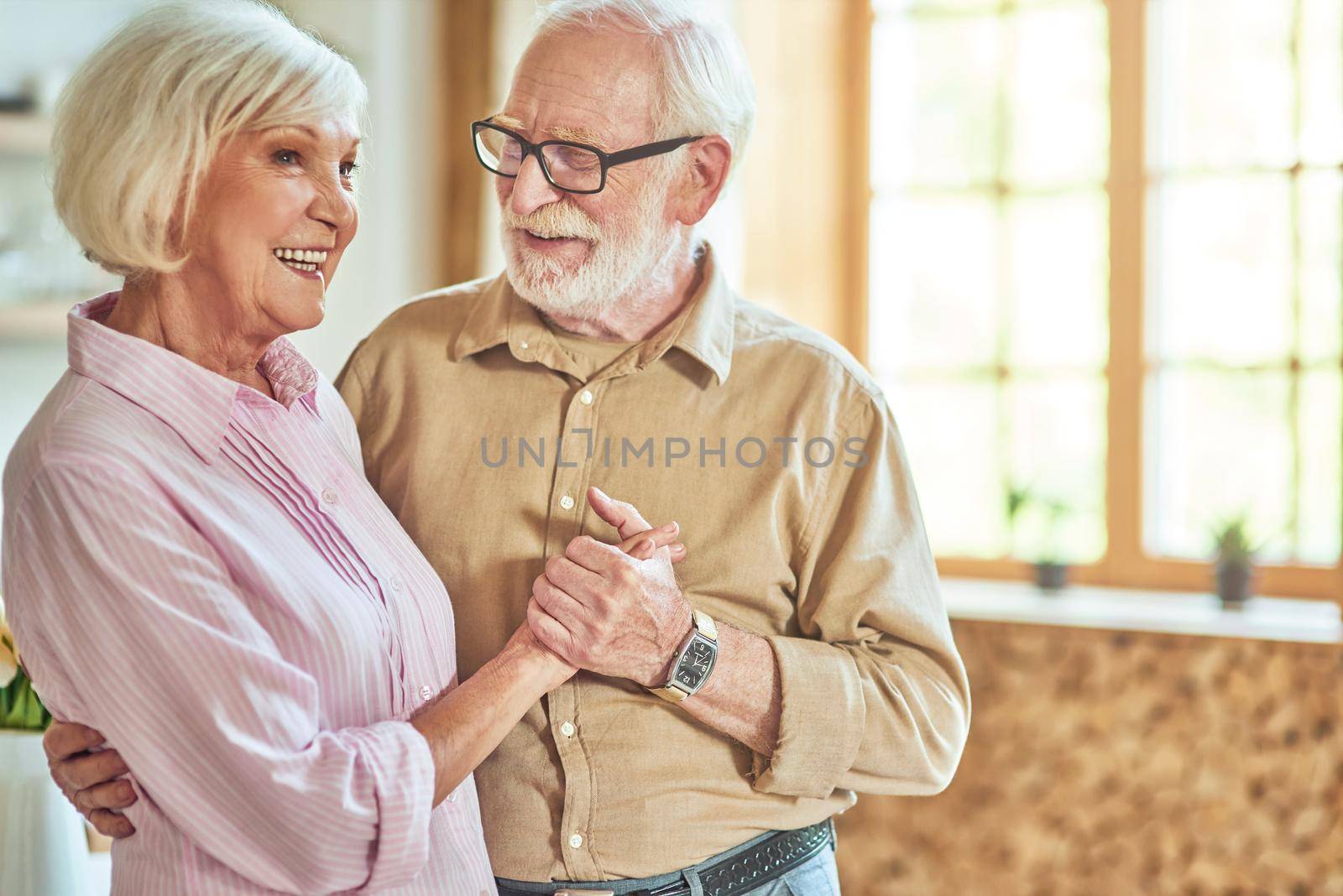 Happy senior man in glasses holding his wife hand while looking at her at home with window on the background. Lifestyle concept