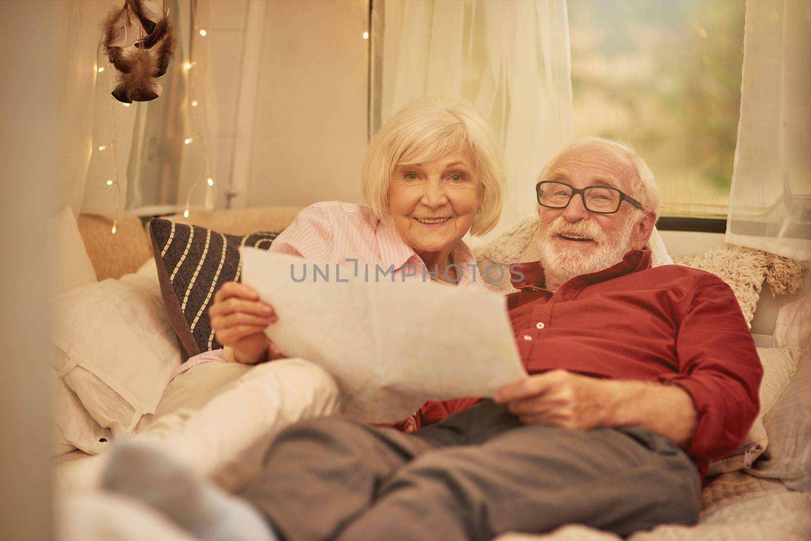 Smiling elderly man and woman lying on couch in camper van while holding map and looking for a new route. Travel concept