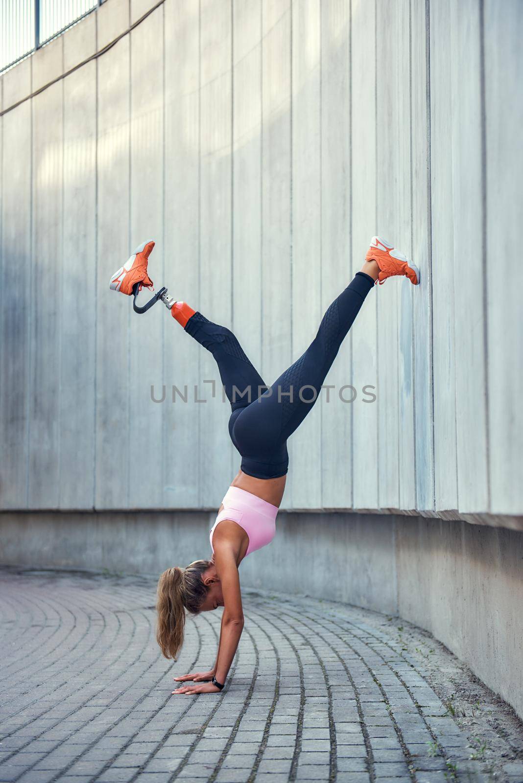 You can do it Full-length of young disabled woman with leg prosthesis in sportswear doing handstand while exercising outdoors. Disabled sport concept by friendsstock