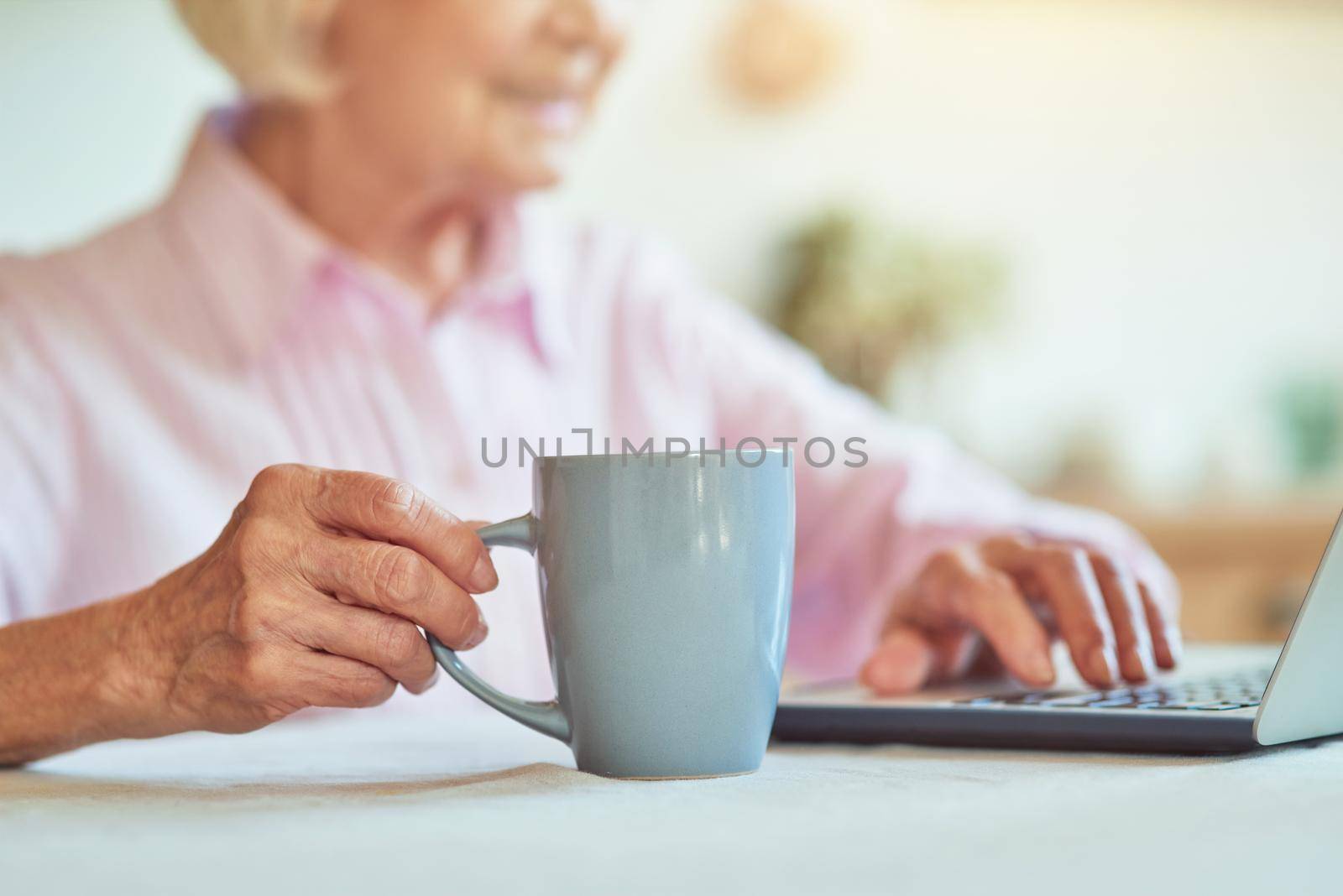 Cropped photo of happy senior woman typing on laptop while enjoying hot drink at home. Domestic lifestyle concept