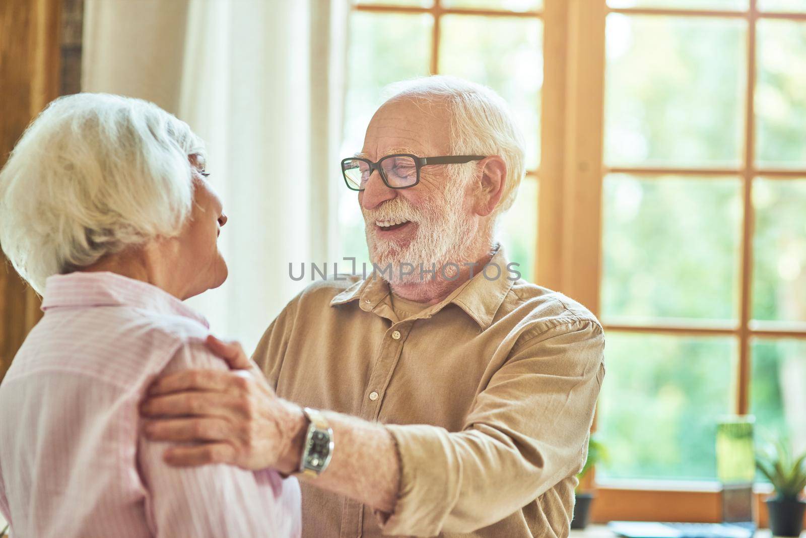 Smiling senior couple looking each other while standing near the window in their house. Family and relationships concept. Copy space