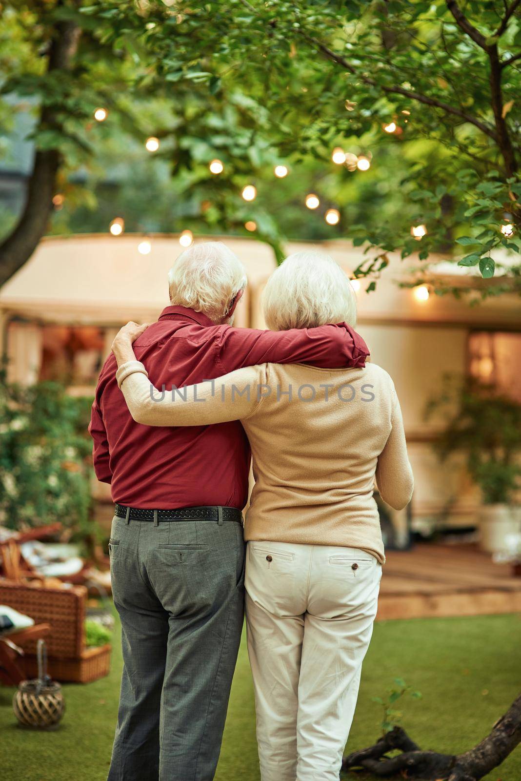 Back view of senior married couple looking at their campervan and hugging while standing outdoors. Travel concept