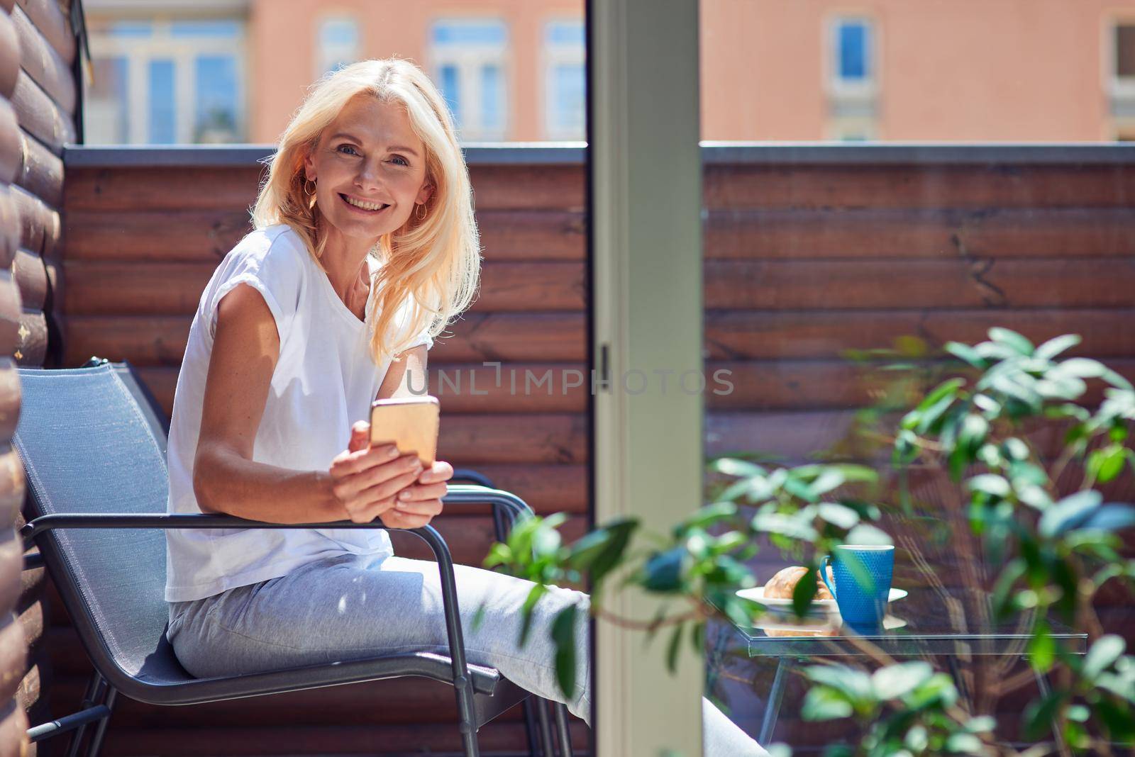 Beautiful middle aged caucasian woman sitting on sunny balcony with phone and smiling at camera. People spending time at home