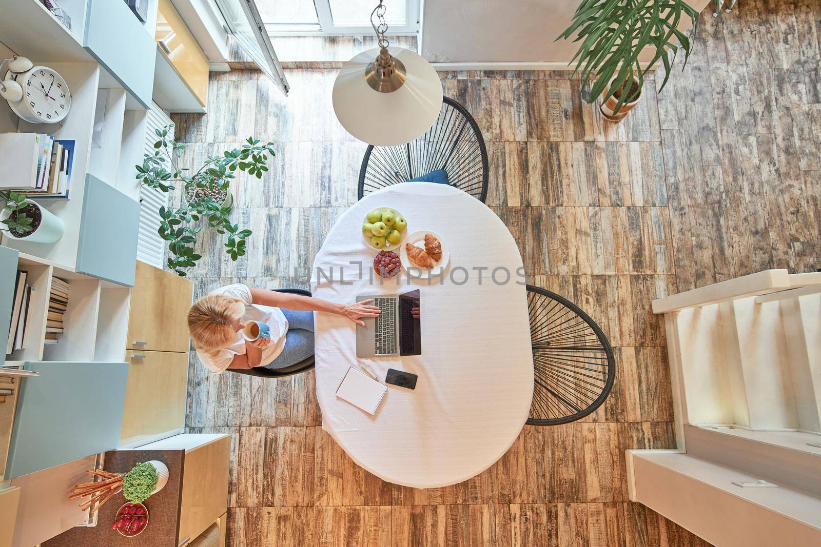 Top view on blonde caucasian business woman drinking coffee while working at home sitting near kitchen table with laptop and smartphone on it. Working from home concept