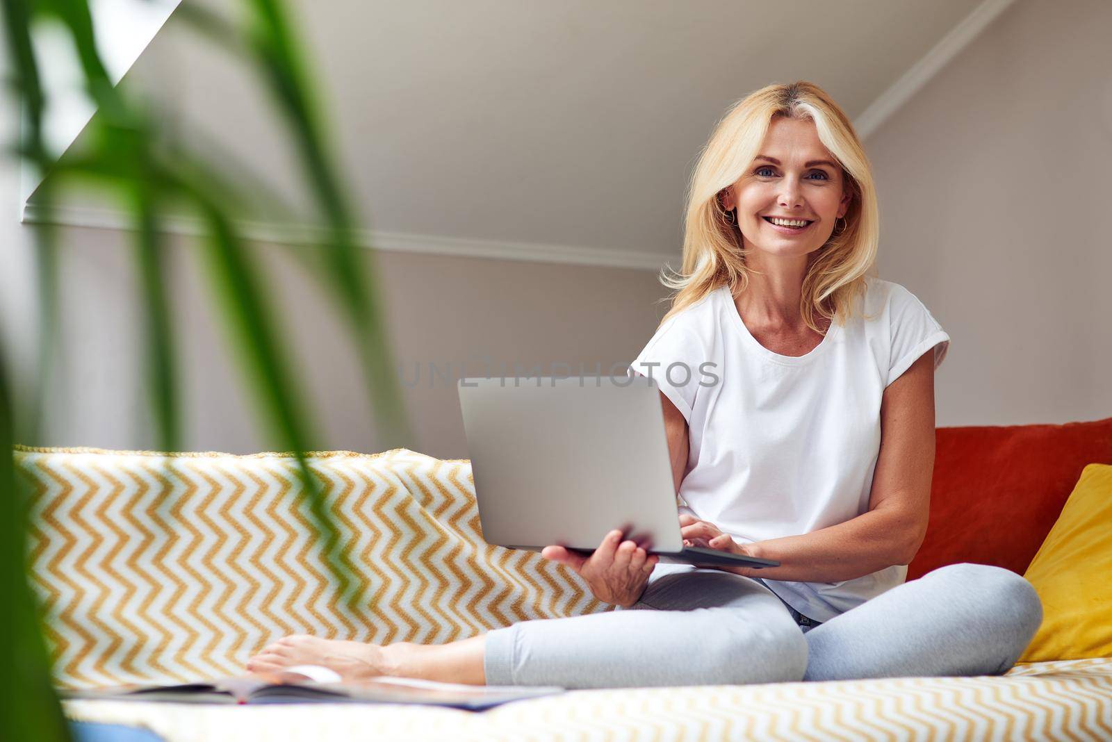 Happy middle aged caucasian woman sitting on sofa with laptop in hands and looking at camera. Working from home concept