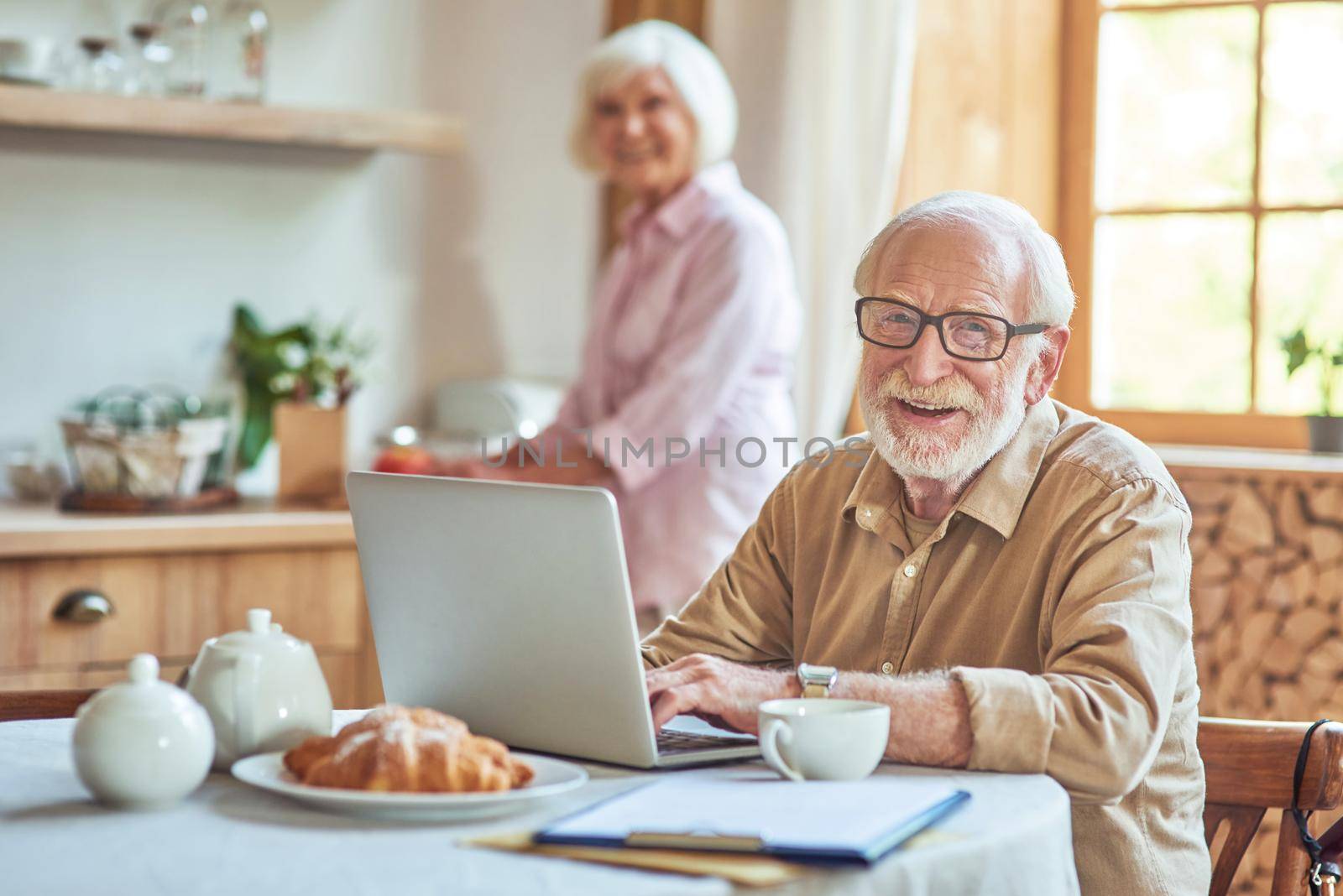 Happy senior man working on laptop while his wife cooking breakfast in the background. Domestic lifestyle concept