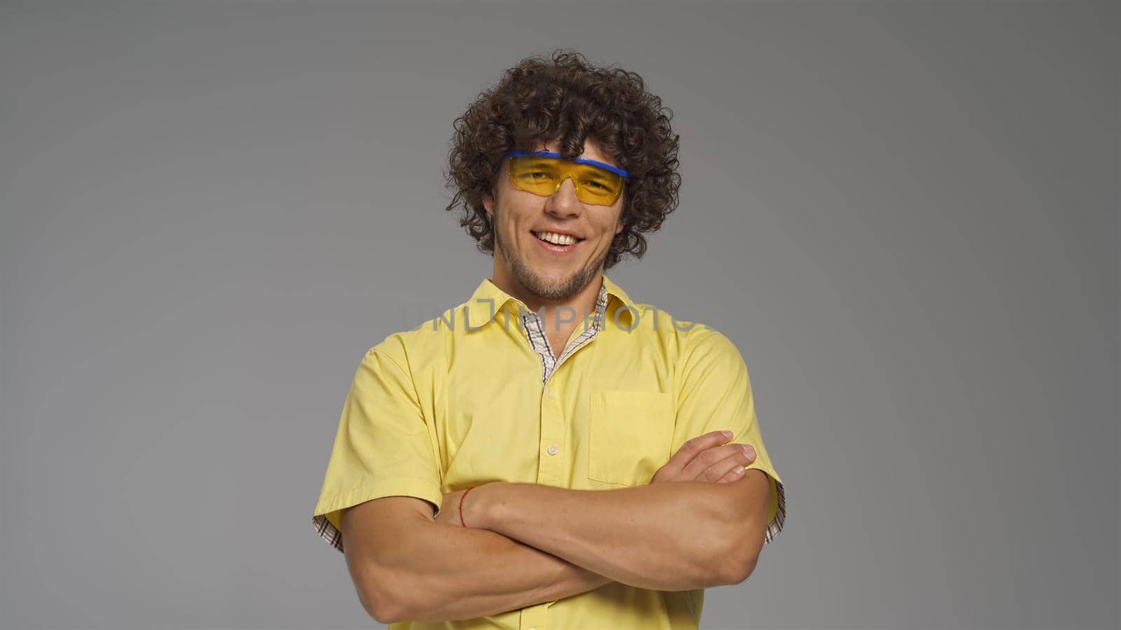 Happy muscular handsome man in goggles smiles with his arms folded. Confident professional on gray background.