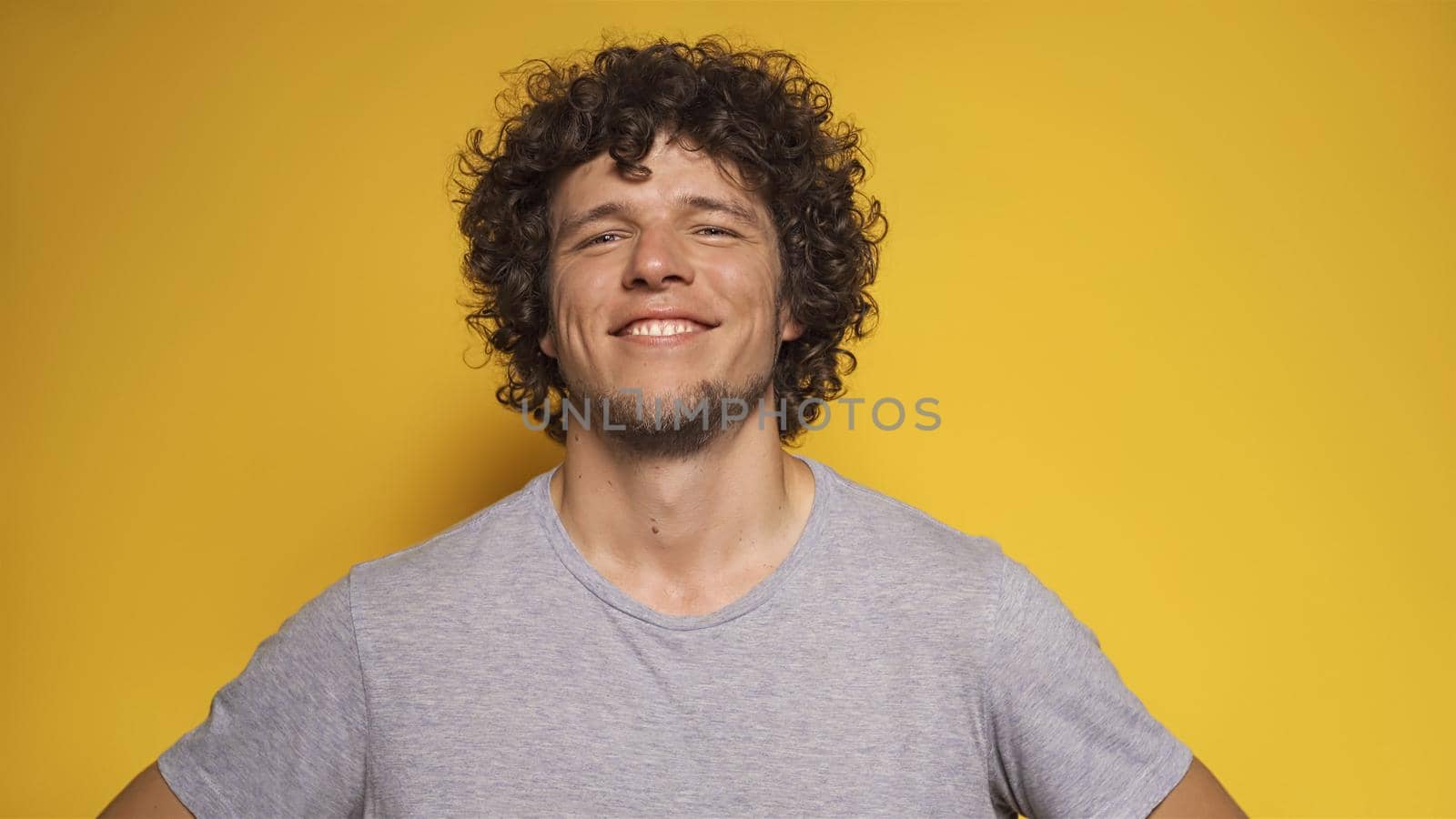Happy young man smiles broadly into camera. Portrait of confident handsome guy with curly or wavy hair on yellow background.