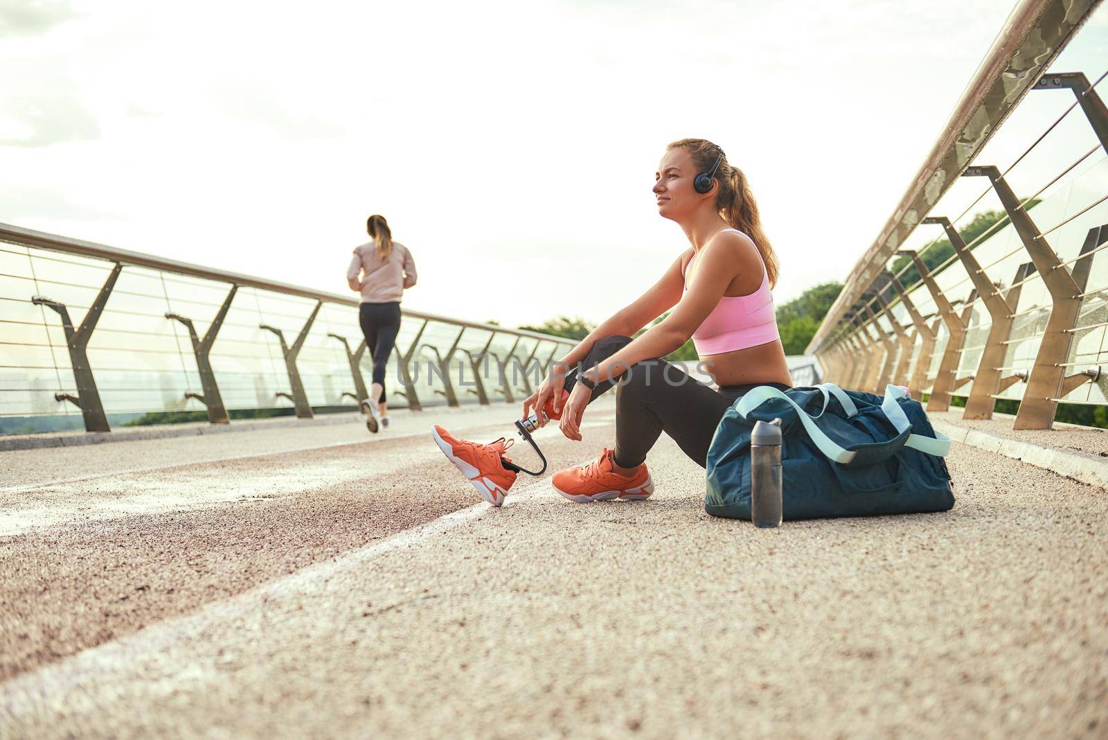 Time to relax. Side view of young beautiful woman in headphones with leg prosthesis listening music and looking aside with smile while sitting on the bridge. Disabled sport concept. Motivation. Healthy lifestyle