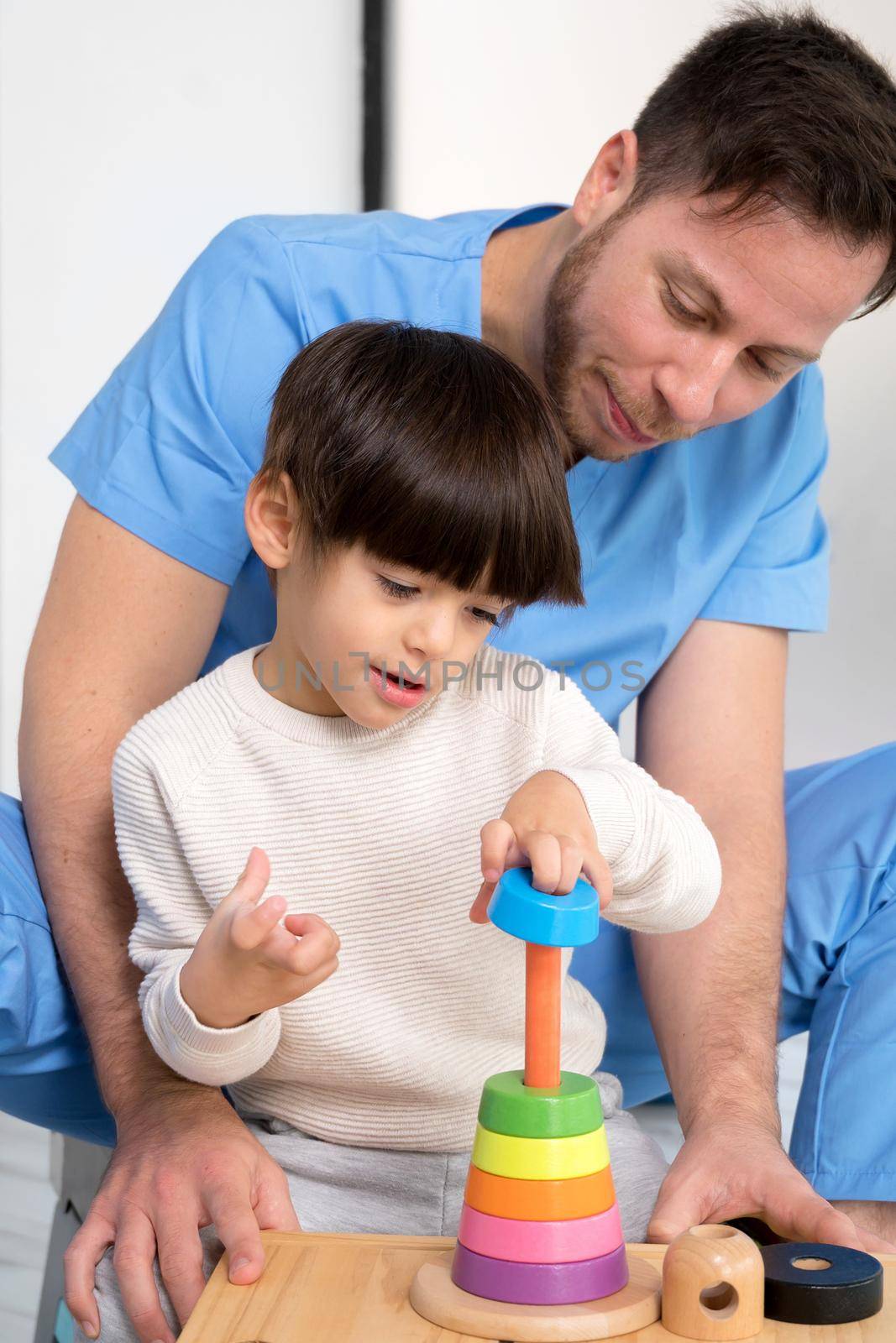 Young therapist helping cute little boy who has cerebral palsy, playing with developing toy at rehabilitation clinic. by HERRAEZ
