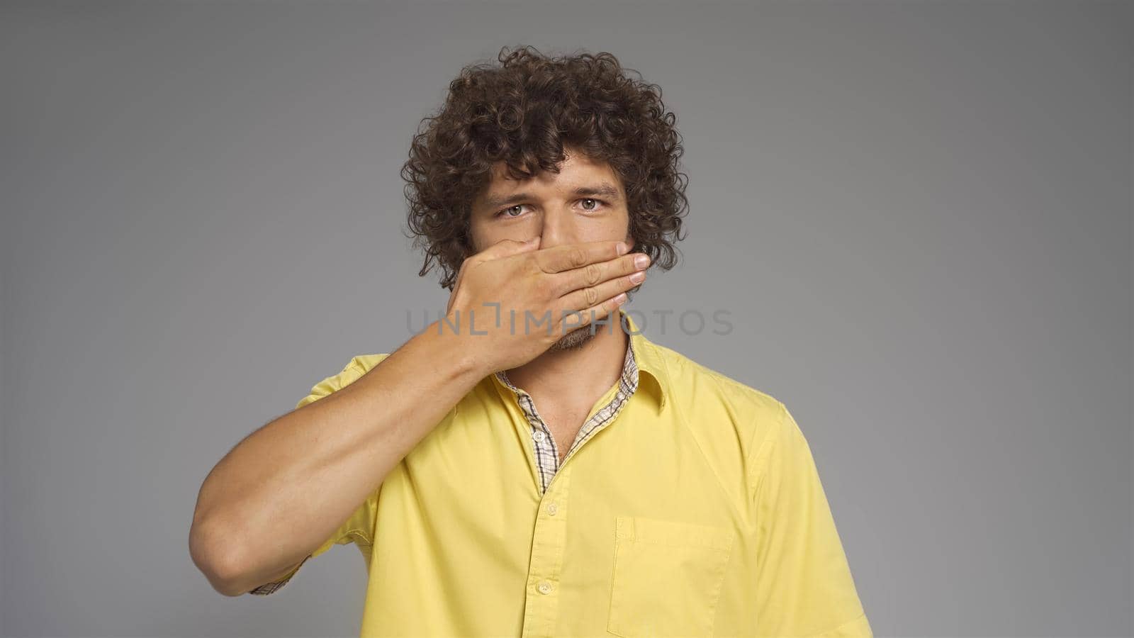 Young Caucasian man covered his mouth with hand looking at camera. Isolated on gray background. Silence concept.
