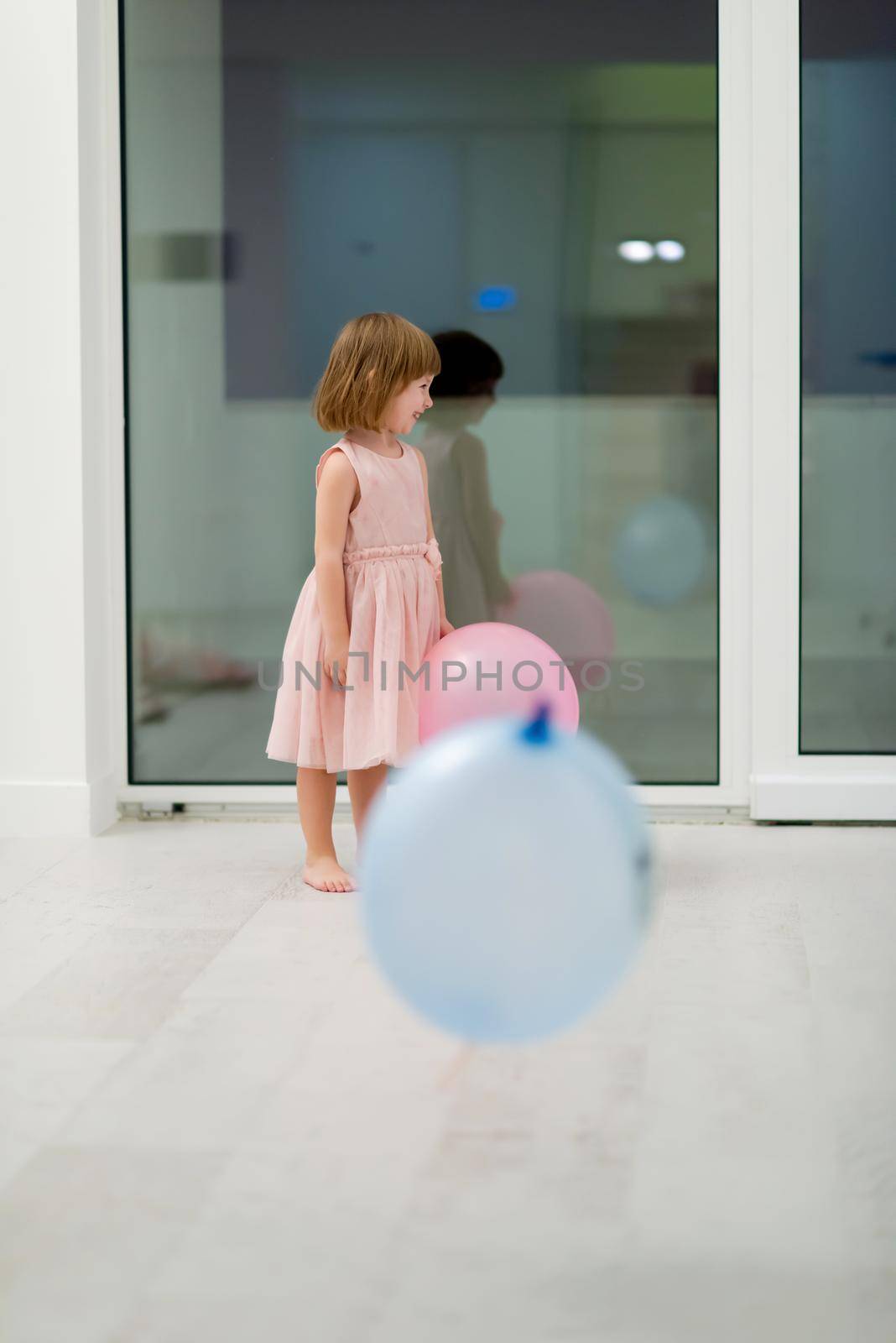 happy cute little girl in a pink dress enjoying while playing with balloons on beautiful evening near the window at home