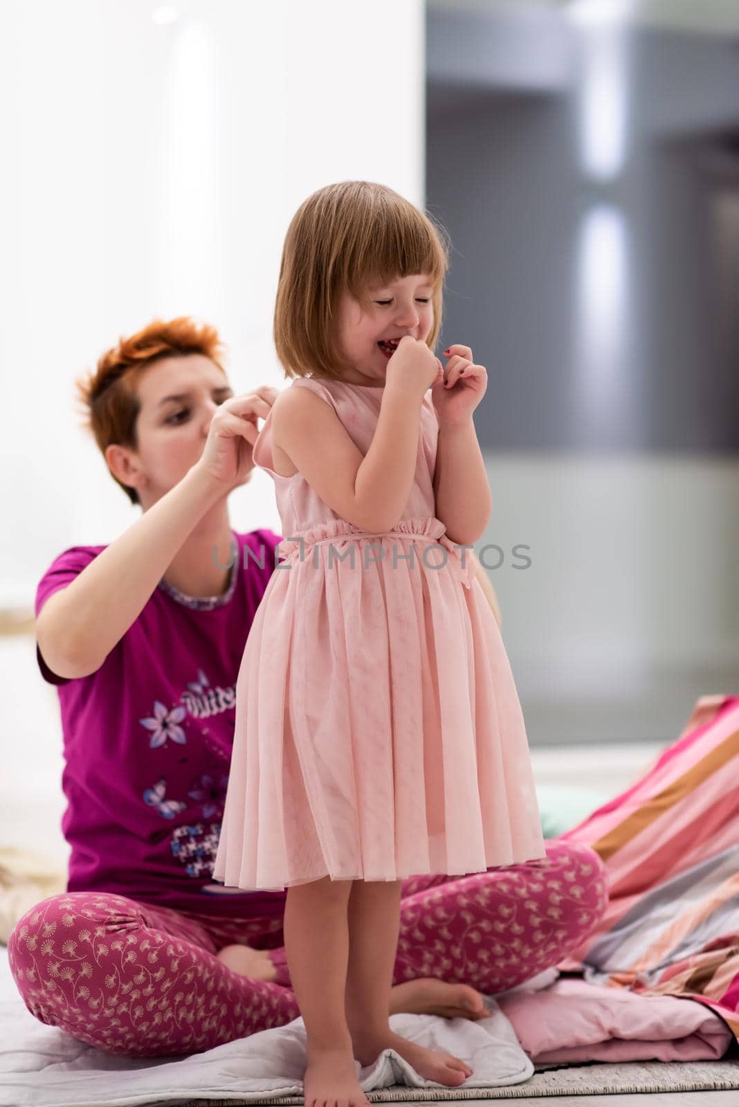 family having fun on beutiful evening at home young mother helping cute little daughter while putting on a pink dress for dancing at home