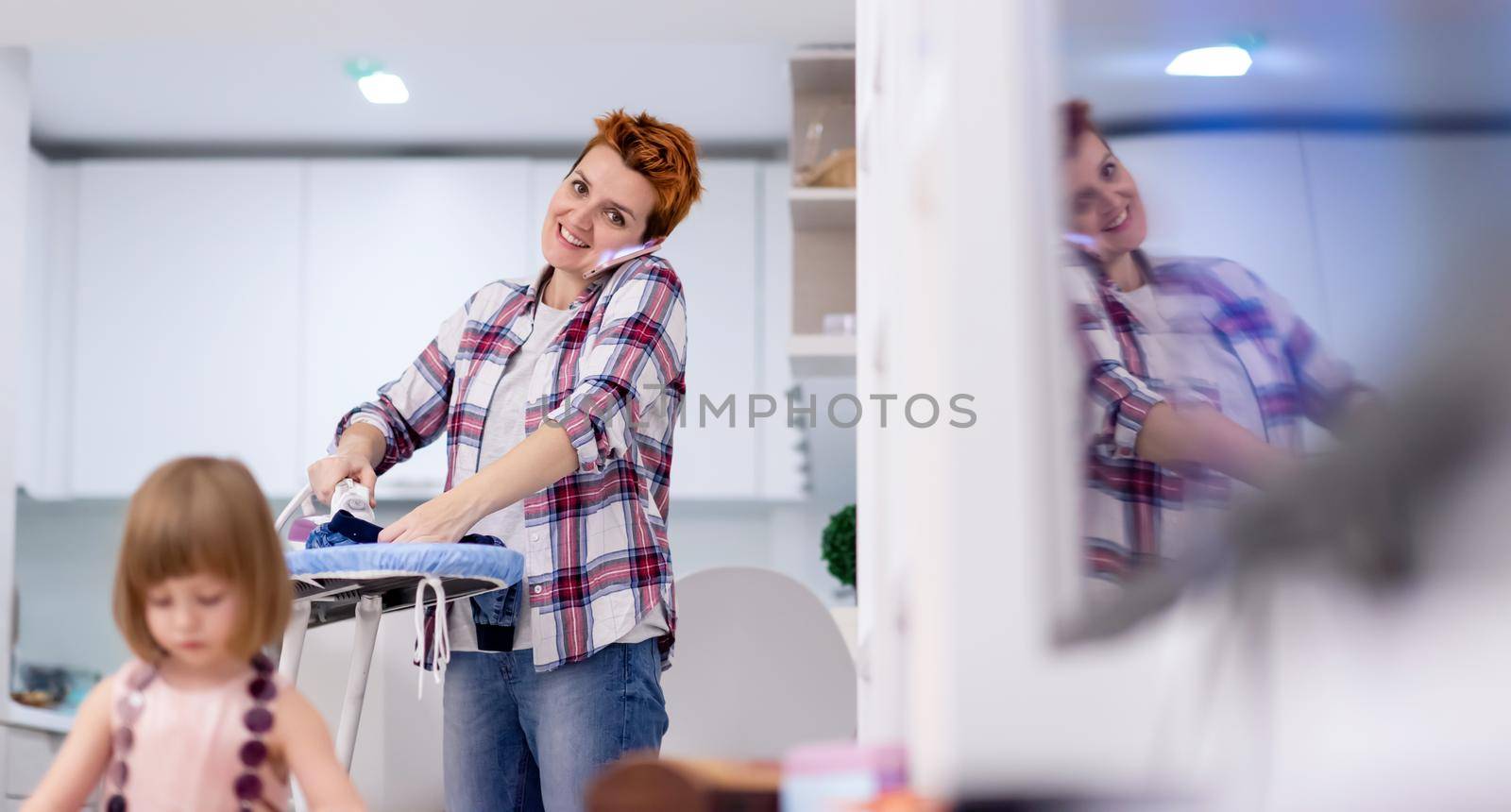 happy family spending time together at home  cute little daughter in a pink dress playing and painting the jewelry box while young redhead mother ironing clothes behind her