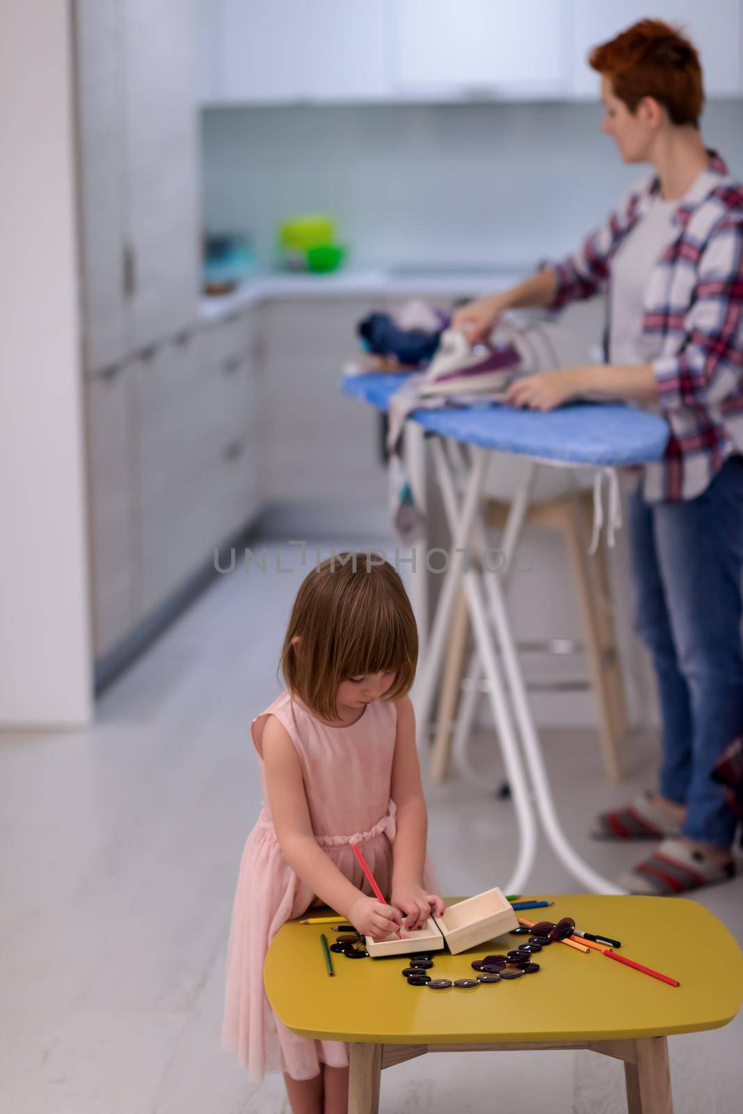 happy family spending time together at home  cute little daughter in a pink dress playing and painting the jewelry box while young redhead mother ironing clothes behind her