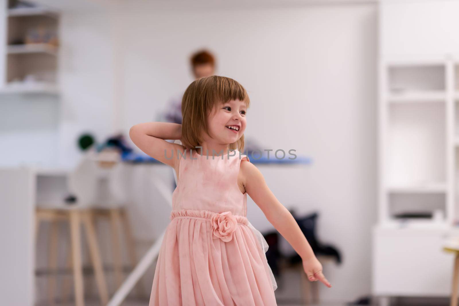 happy family having fun together at home  cute little daughter in a pink dress playing and dancing while young redhead mother ironing clothes behind her