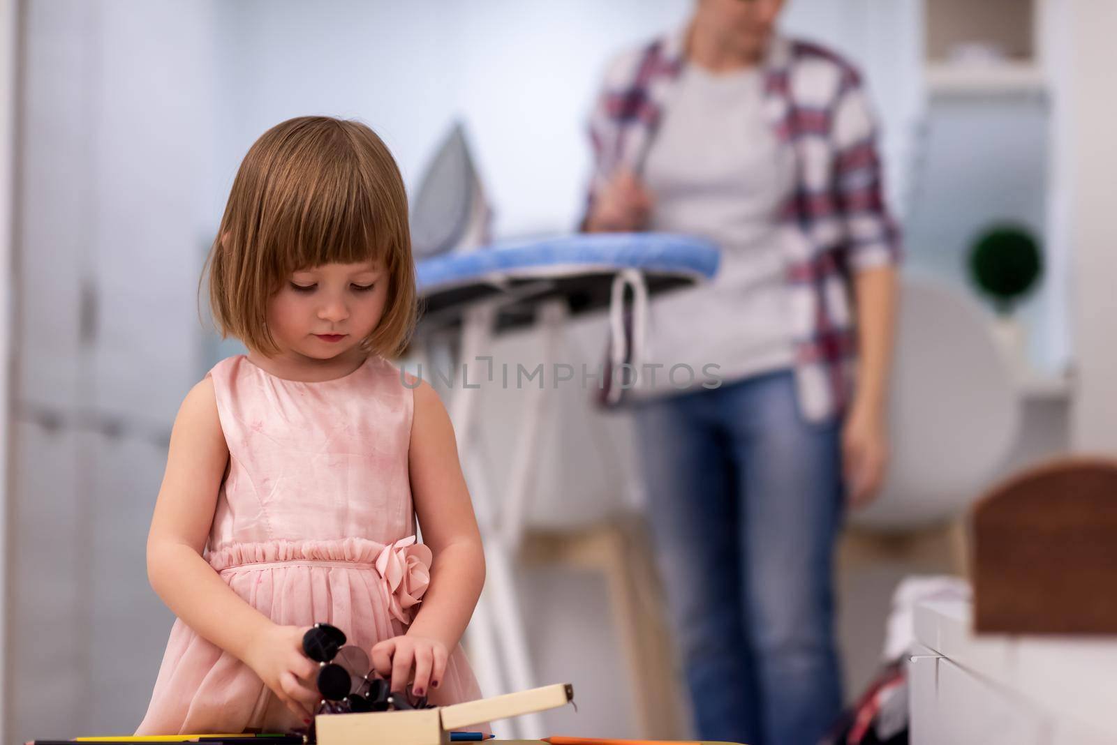 happy family spending time together at home  cute little daughter in a pink dress playing and painting the jewelry box while young redhead mother ironing clothes behind her