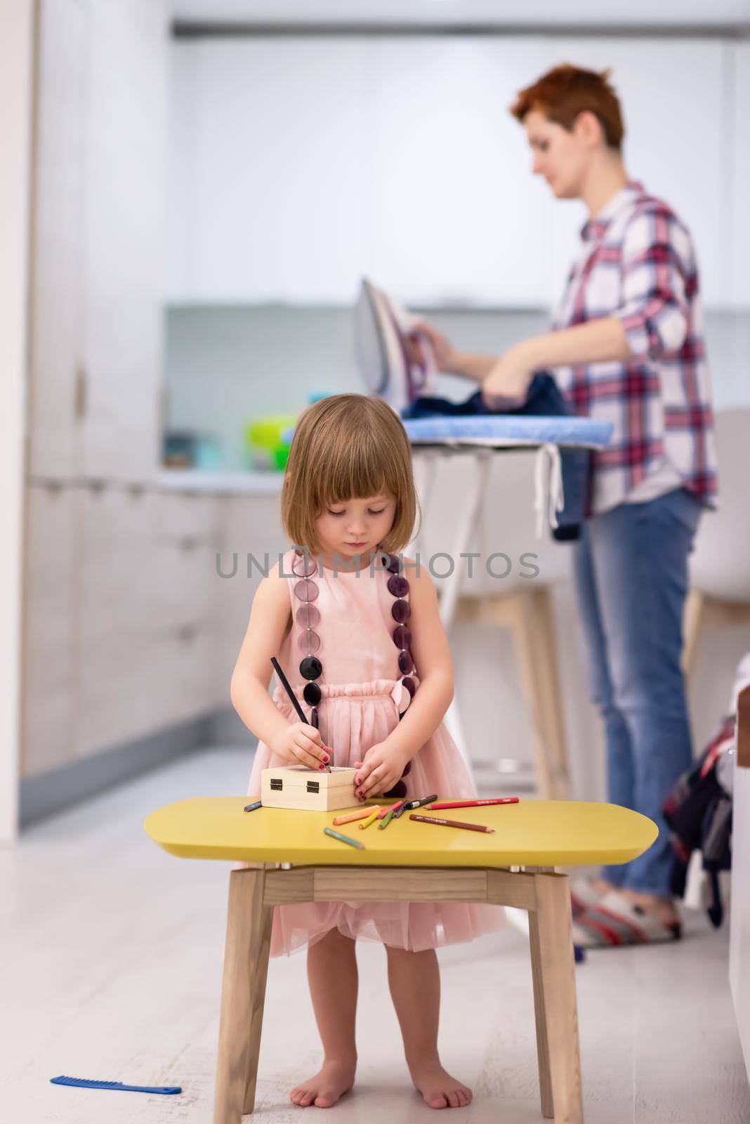 happy family spending time together at home  cute little daughter in a pink dress playing and painting the jewelry box while young redhead mother ironing clothes behind her