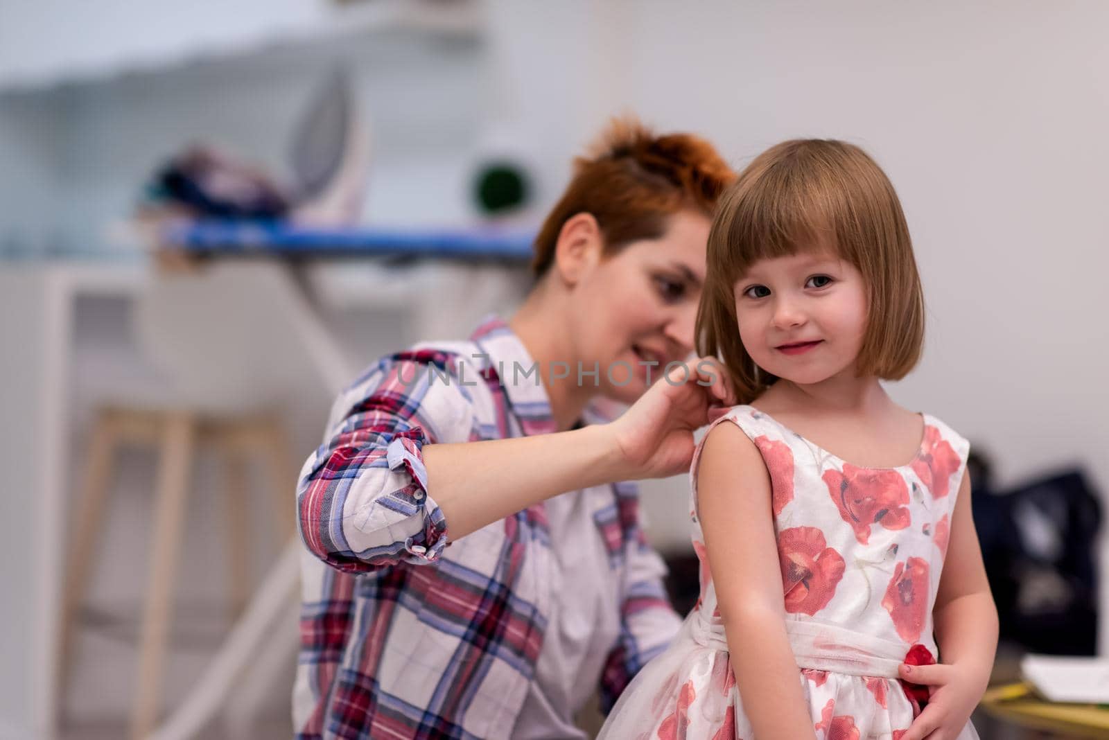 family having fut on beautiful evening  young mother helping cute little daughter while putting on a dress with red flowers for dancing at home