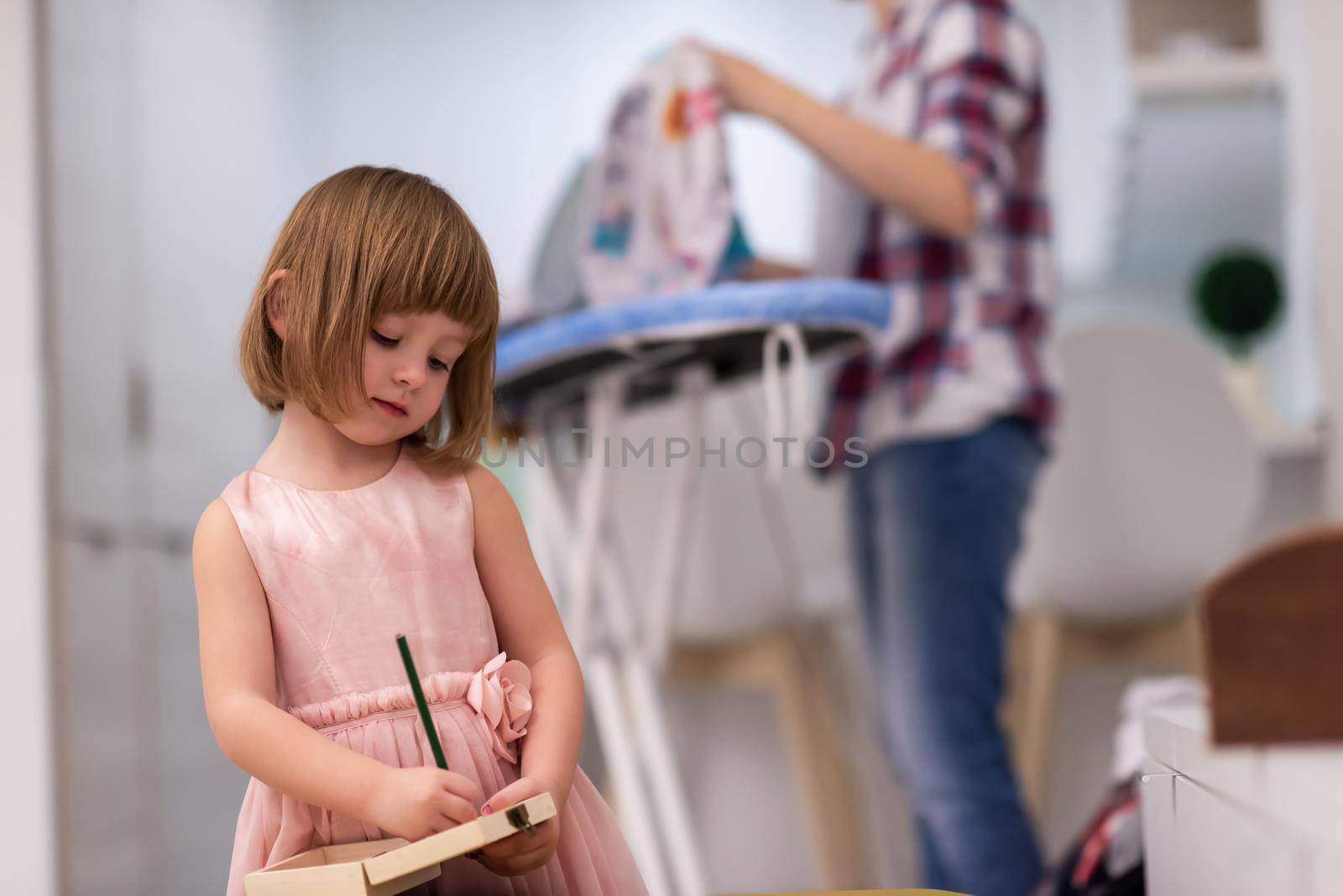 happy family spending time together at home  cute little daughter in a pink dress playing and painting the jewelry box while young redhead mother ironing clothes behind her