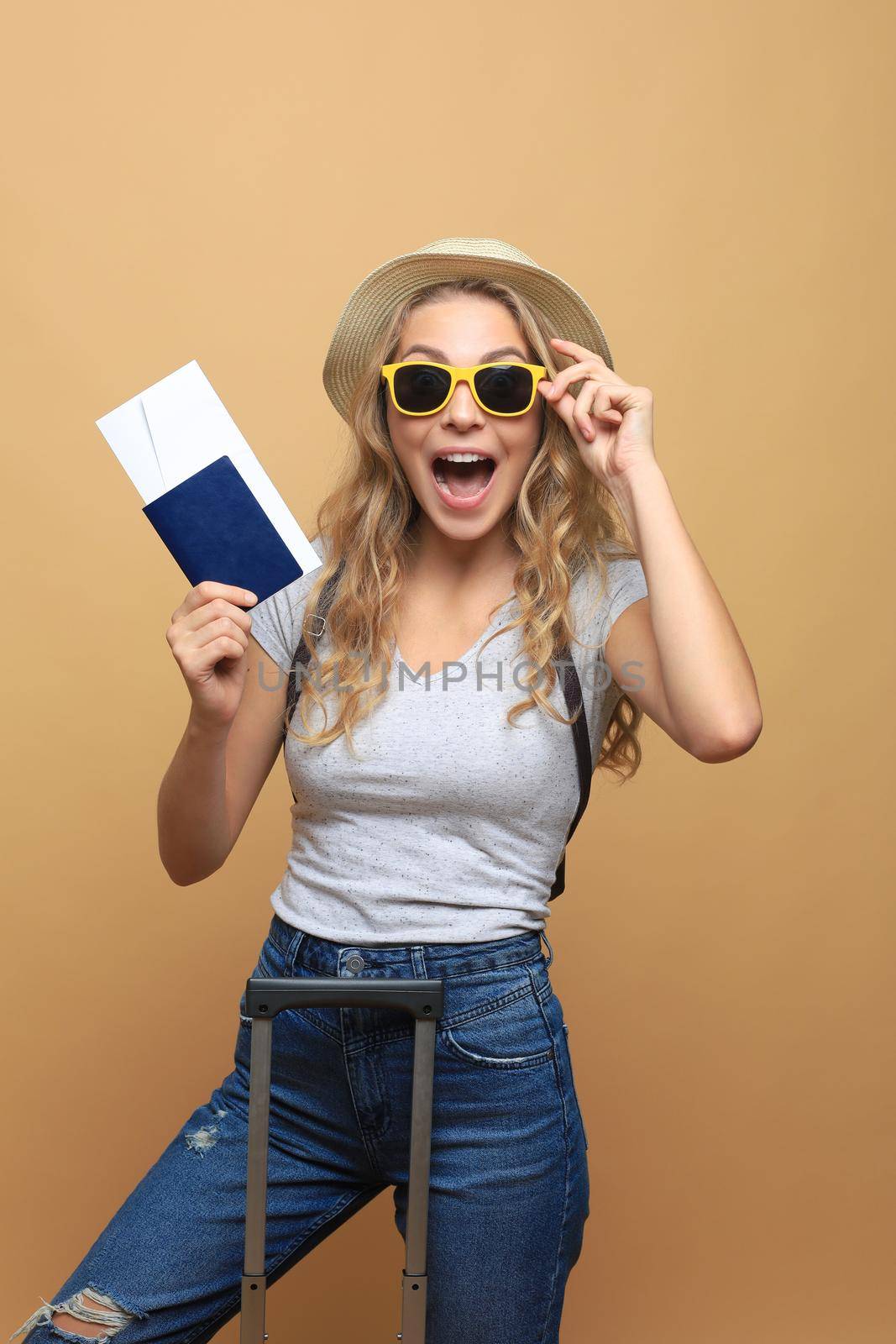 Cheerful blonde woman posing with baggage and holding passport with tickets while looking away over beige background. by tsyhun