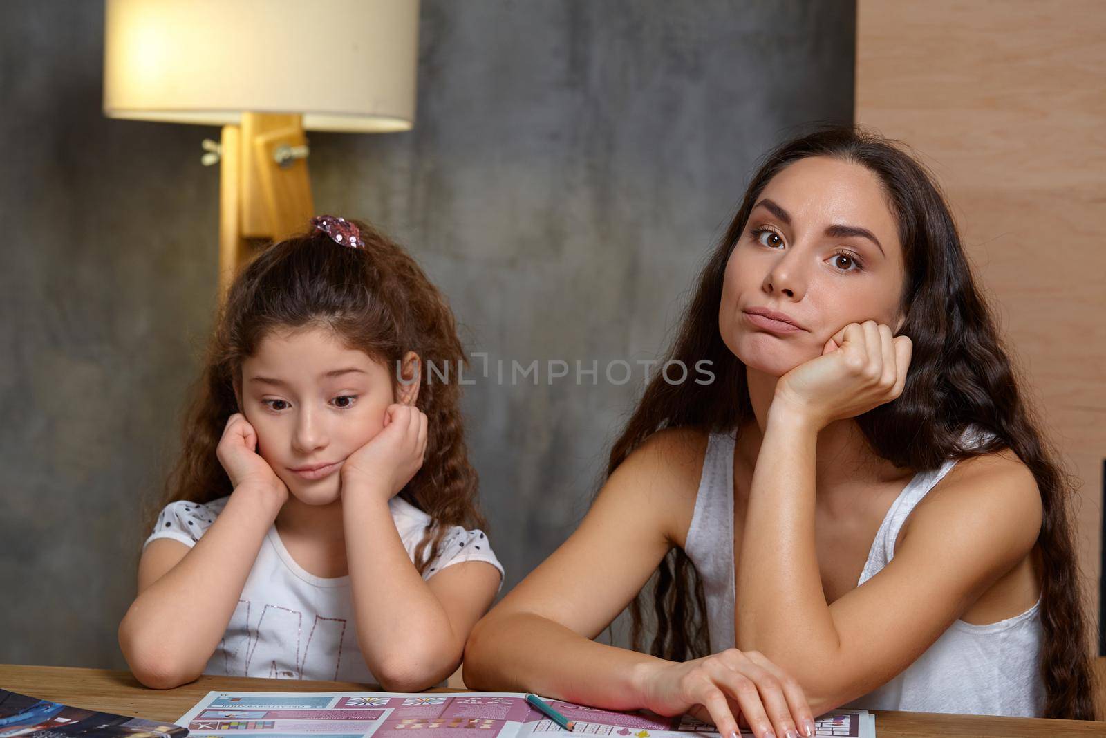 Portrait of a mother helping her small sweet and cute daughter to make her homework indoors, propping up their heads.