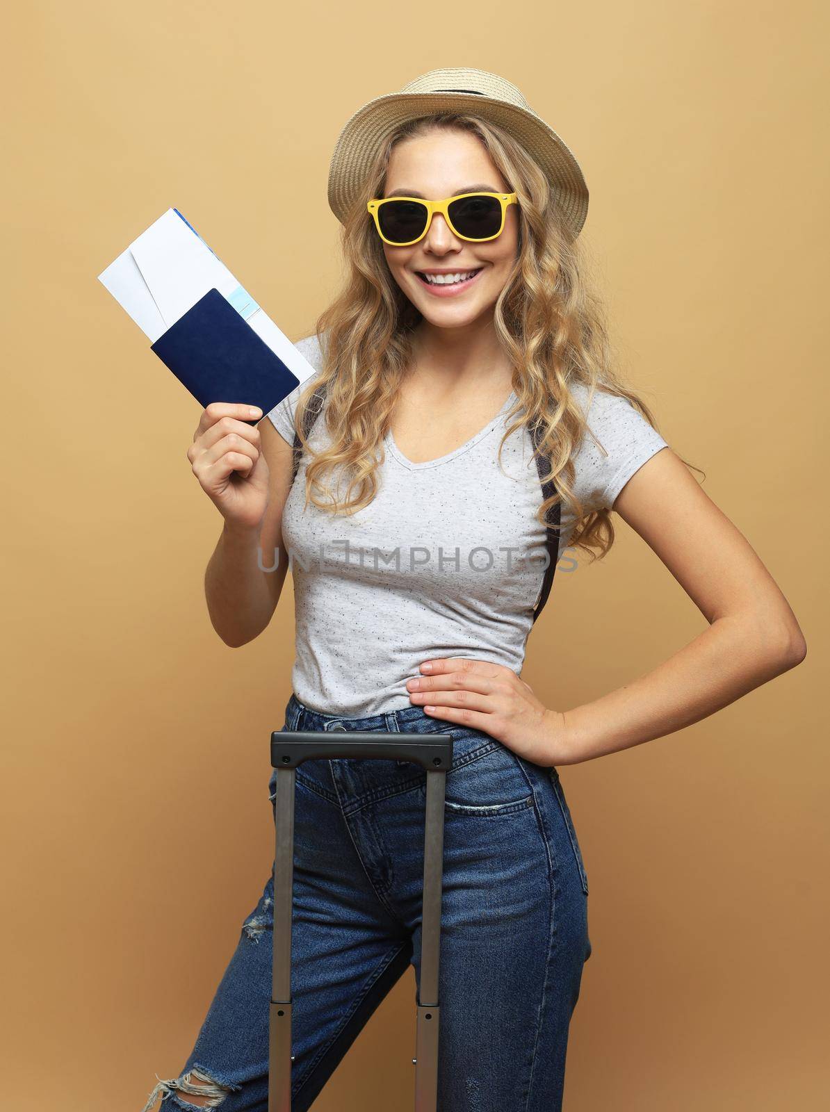 Cheerful blonde woman in sunglasses posing with baggage and holding passport with tickets over beige background